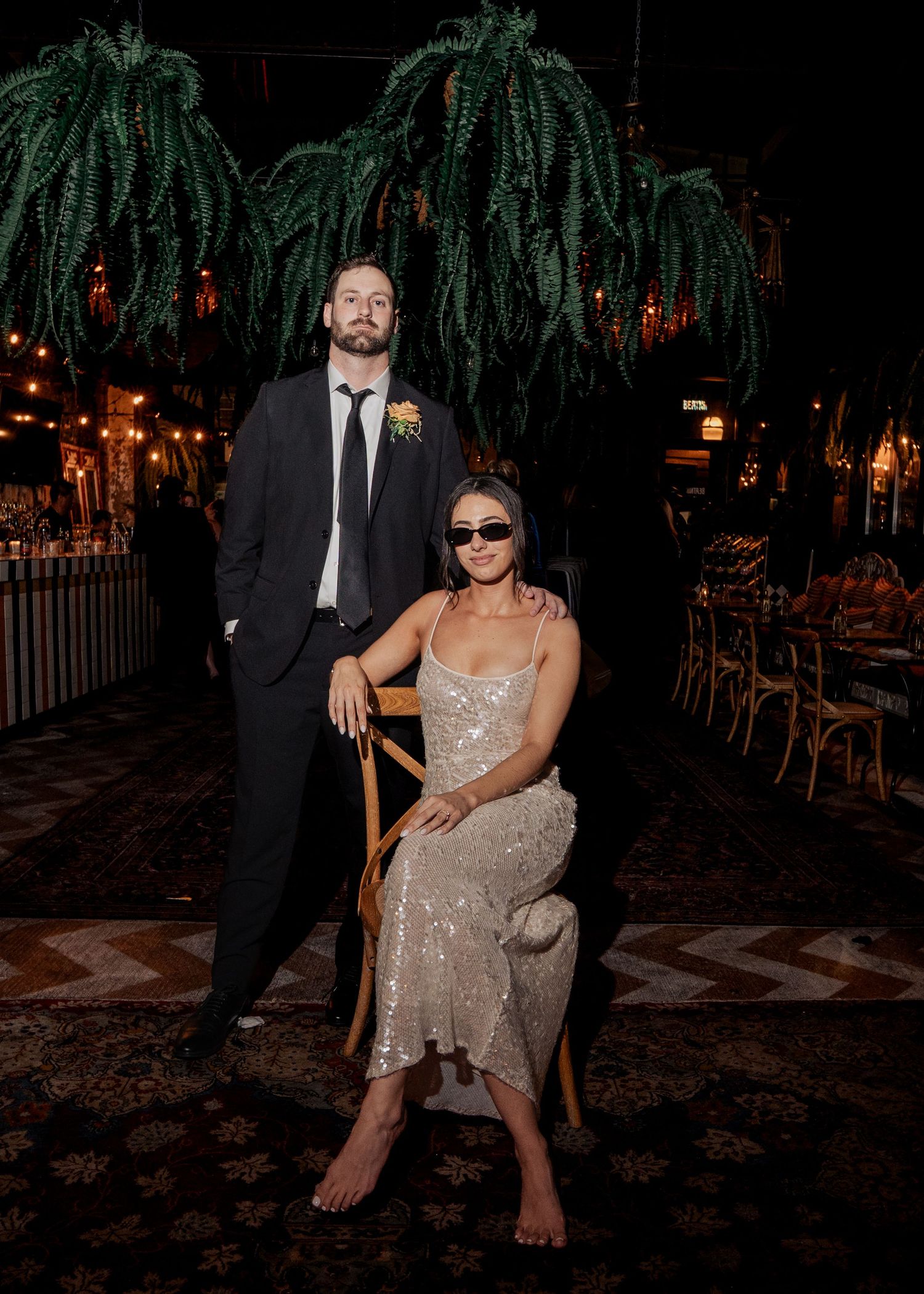 Couple posing at night wedding reception, man in black suit standing behind woman in sequined dress wearing sunglasses.