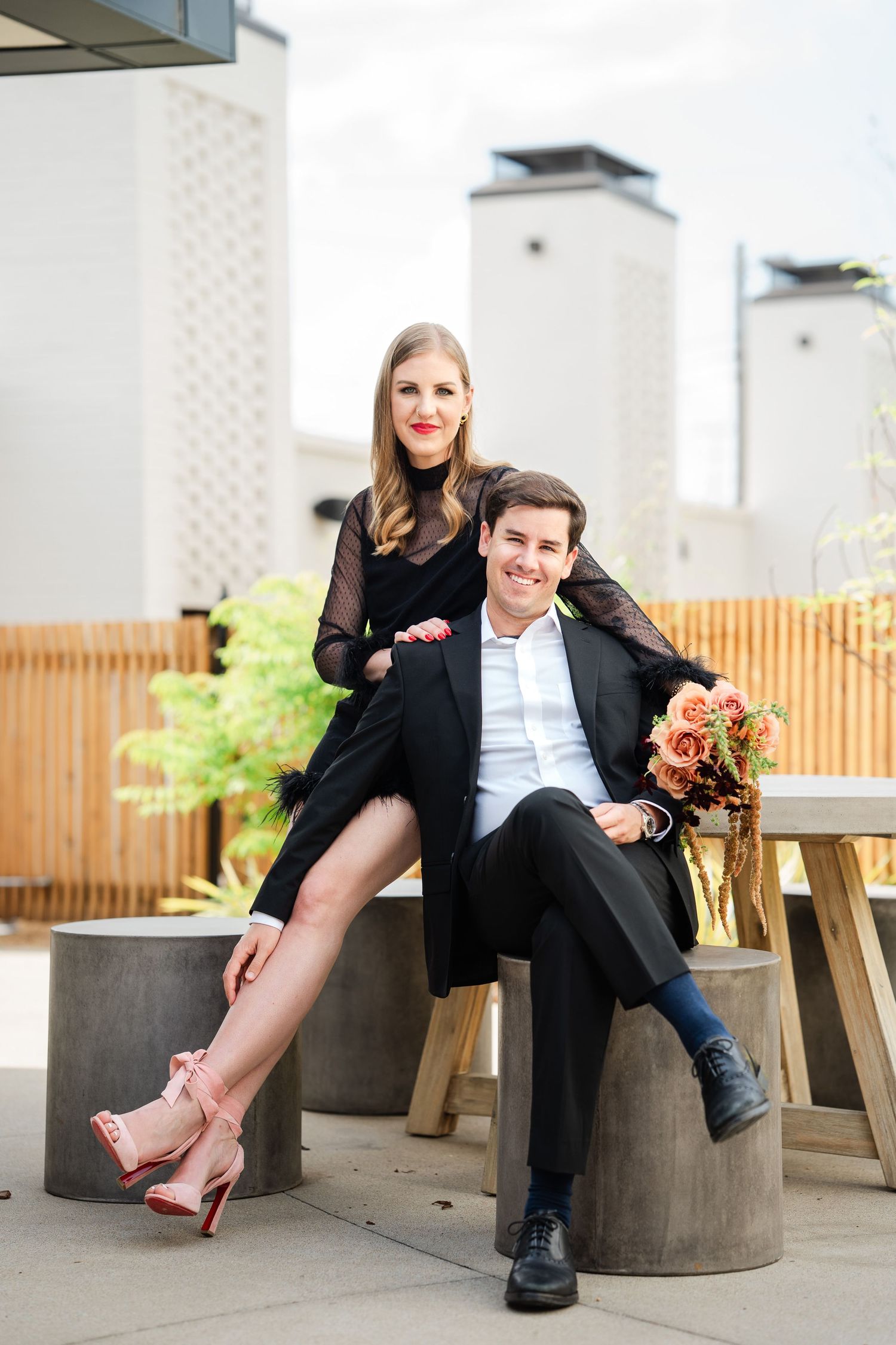 Elegant couple posing on rooftop patio, woman in black dress with pink heels, man in black suit holding floral bouquet.