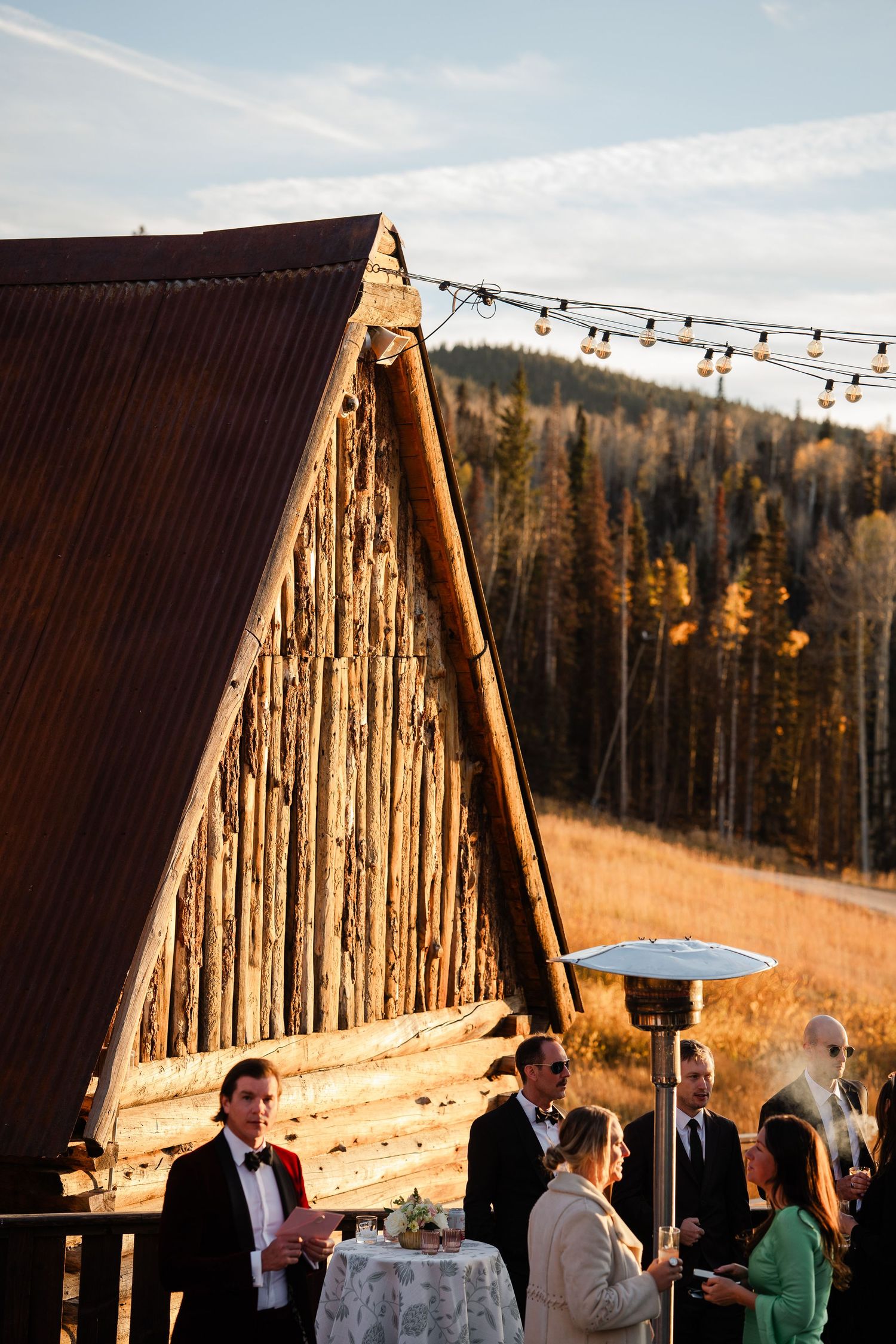 Wedding cocktail hour guests mingling outside rustic A-frame barn with string lights at golden hour.