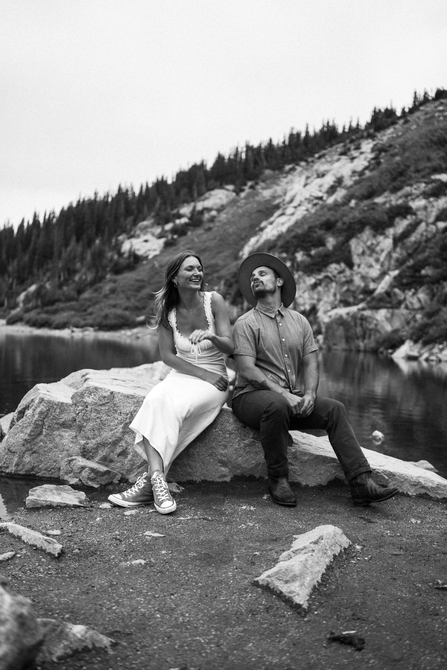 Black and white photo of a couple sitting on rocks by a mountain lake, laughing and enjoying each others company.