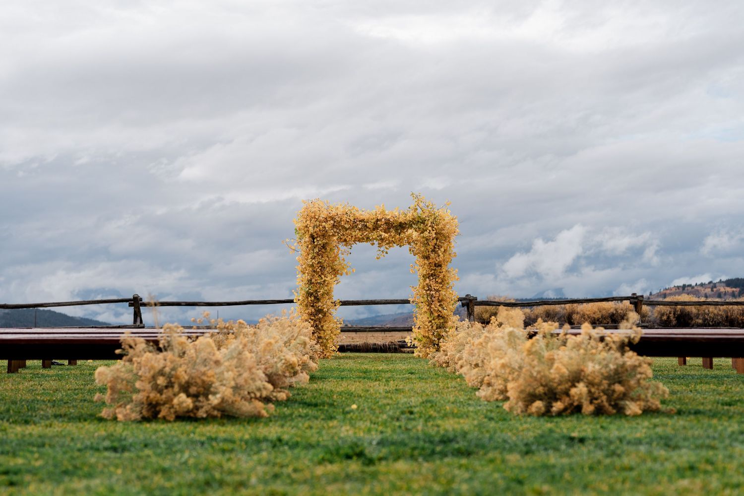 Outdoor wedding arch adorned with golden dried flowers on green grass under a cloudy sky.