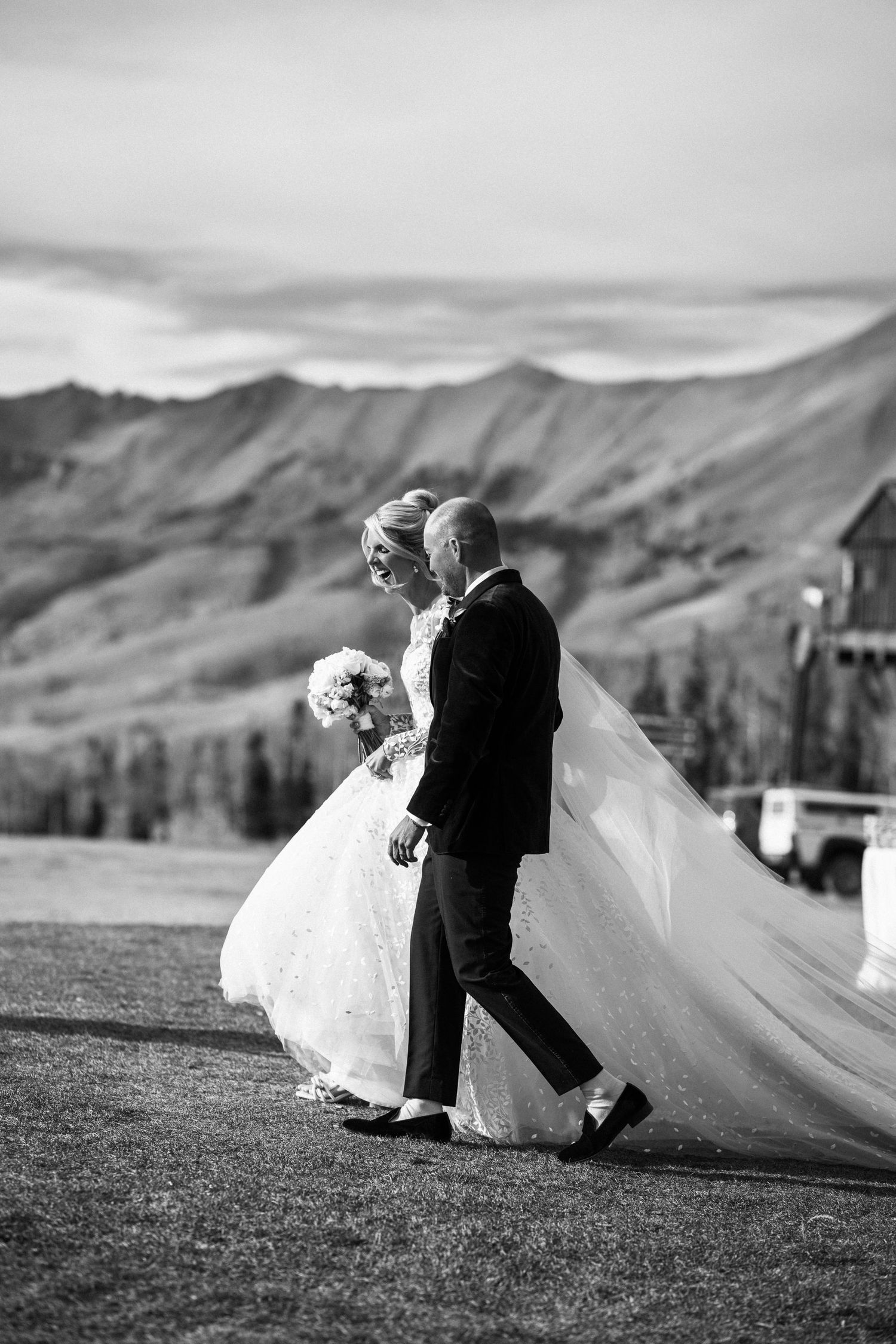 Black and white photo of bride and groom walking together with flowing veil against mountain landscape backdrop.