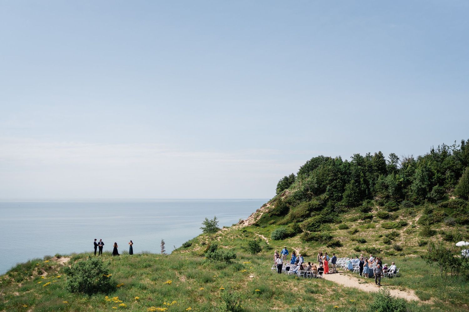 Outdoor wedding ceremony on a scenic coastal cliff with guests gathered among lush green hills overlooking the ocean.