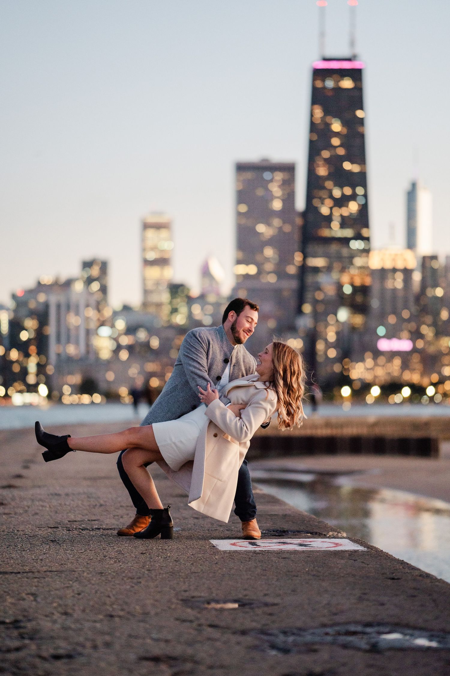 Couple sharing a romantic dip kiss on Chicago waterfront with city skyline bokeh lights at dusk.