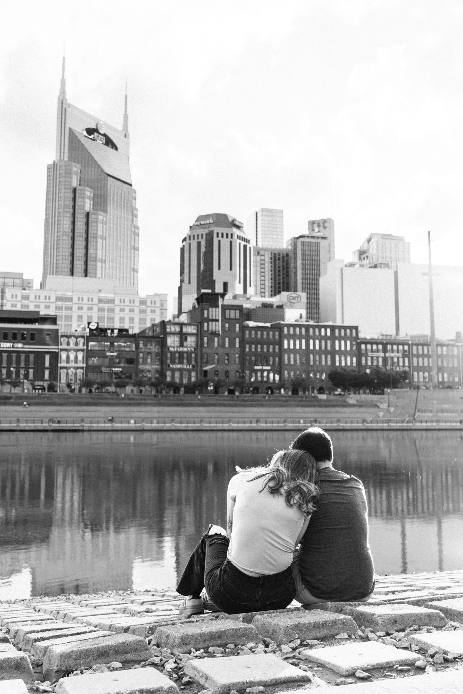 Black and white photo of couple sitting together by a river with Nashville city skyline behind them.