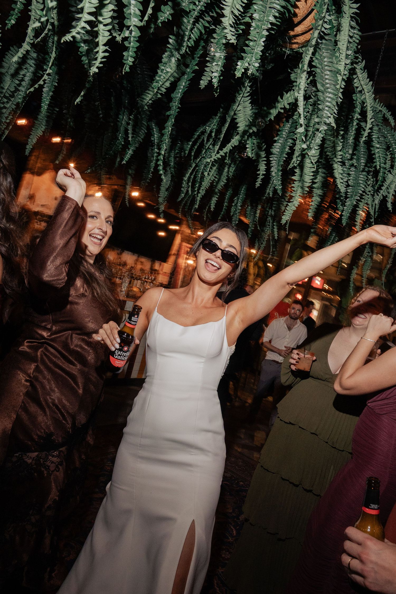 Bride in white gown wearing sunglasses dancing and celebrating with guests under lush hanging ferns at wedding reception.