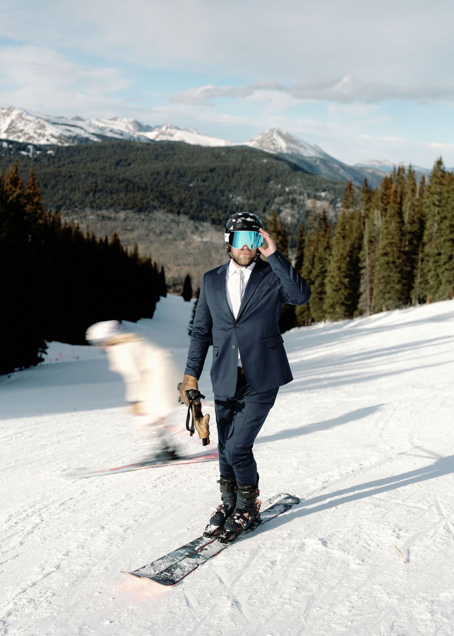 Groom in navy suit adjusts ski goggles on helmet while snowboarder passes on sunny mountain slope.