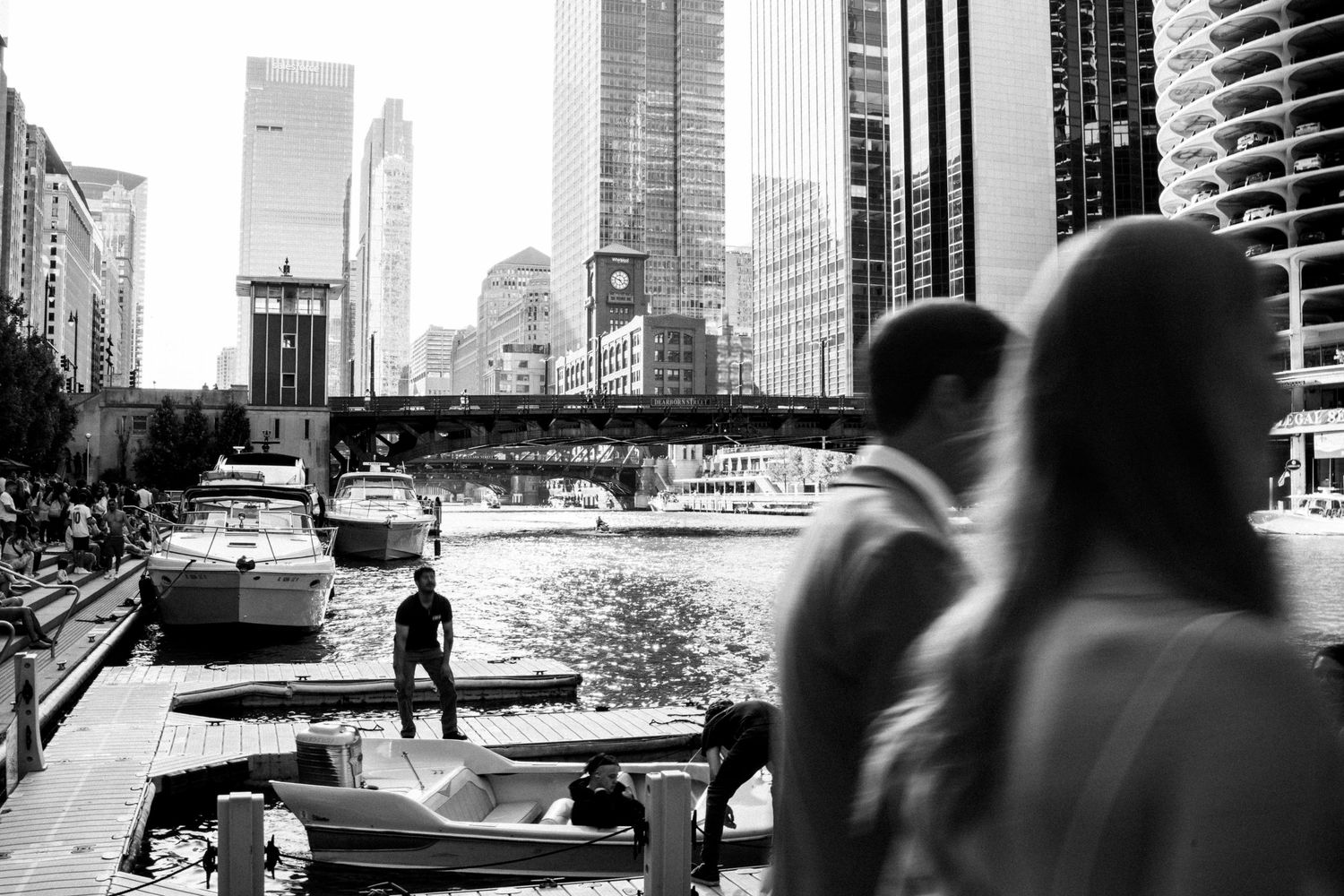 Black and white photo of couple near Chicago Riverwalk with city skyline and boats in background.