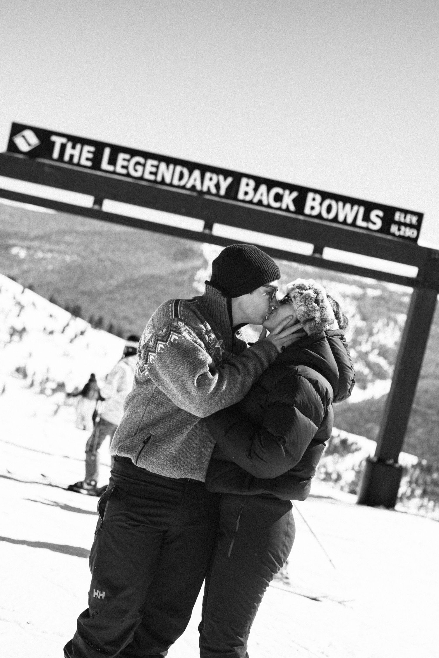 Black and white photo of a couple sharing a kiss under The Legendary Back Bowls sign at a snowy ski resort.
