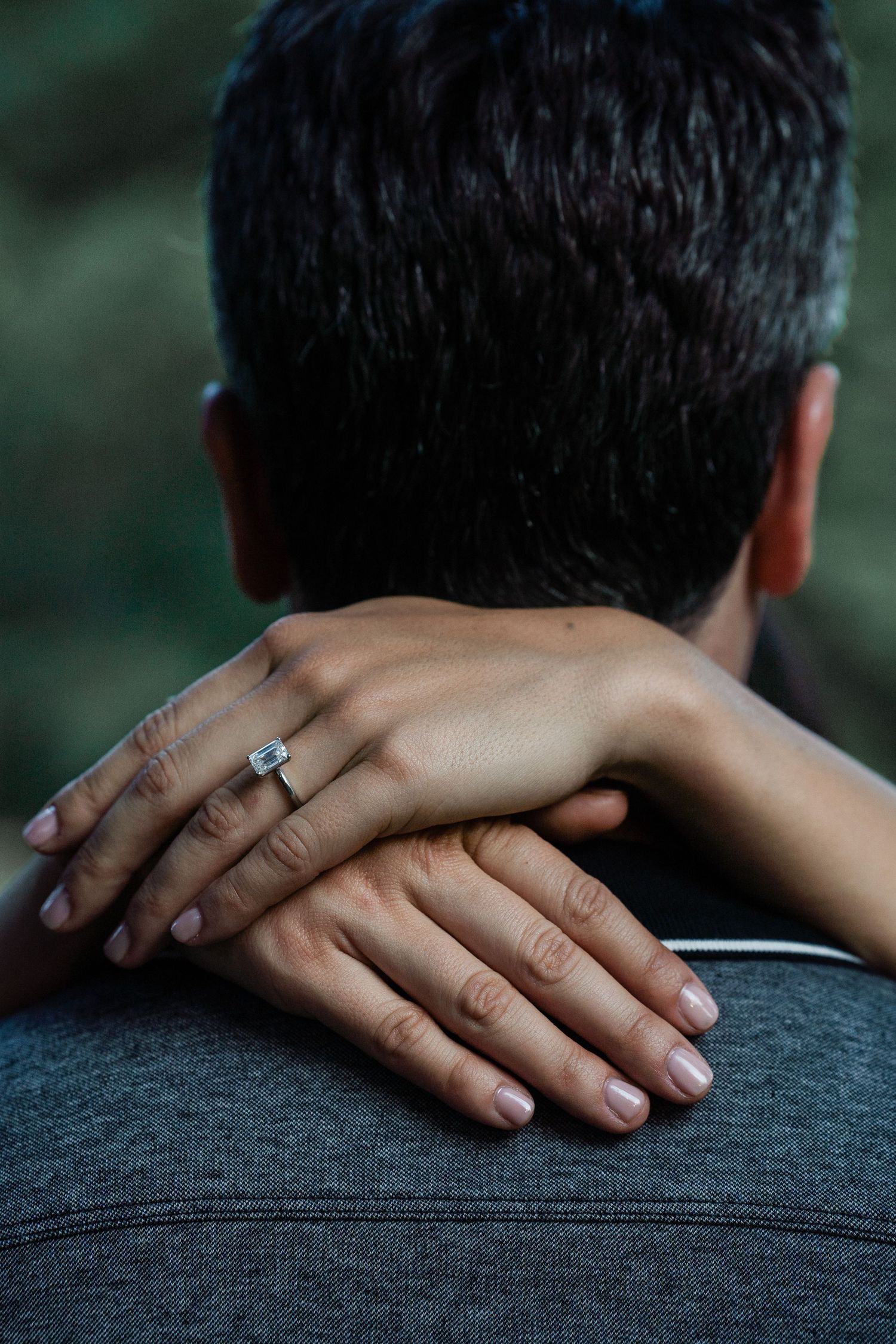 Close-up of woman's hand with emerald cut engagement ring resting on man's shoulder.