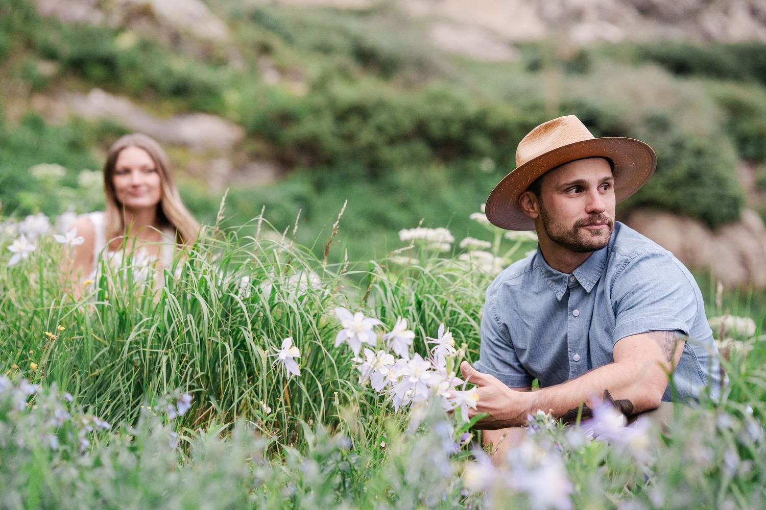 Couple relaxing among white wildflowers in a lush green mountain meadow during engagement photo session.