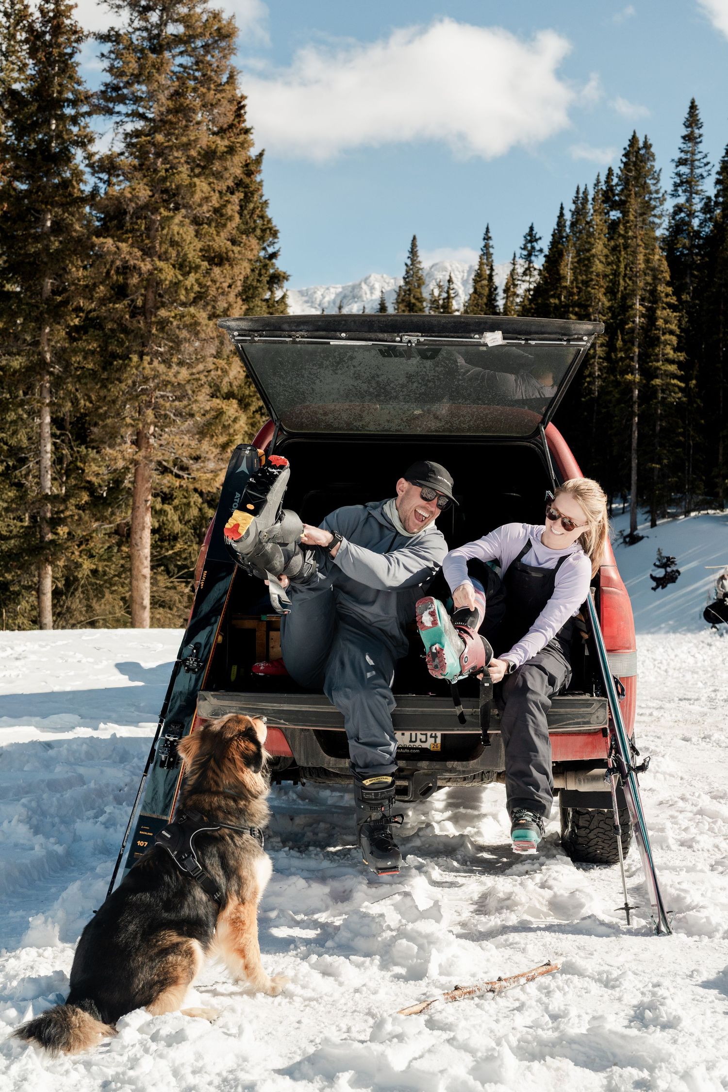 Couple sitting in truck tailgate putting on ski boots with dog waiting in snowy forest.