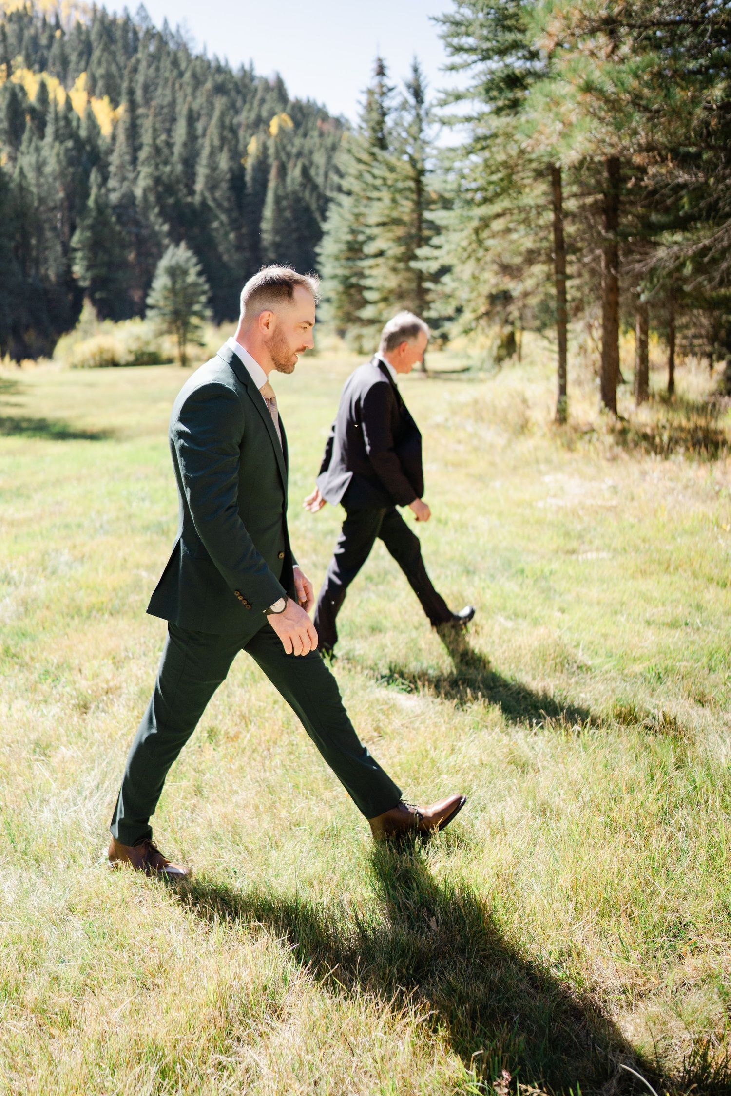 Groom in dark suit walks confidently through a sunny mountain meadow surrounded by pine trees on wedding day.