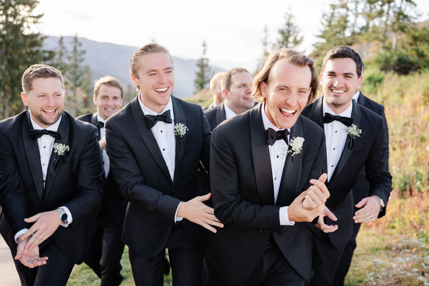 Groomsmen in black tuxedos laughing and celebrating outdoors with mountain views at a wedding.