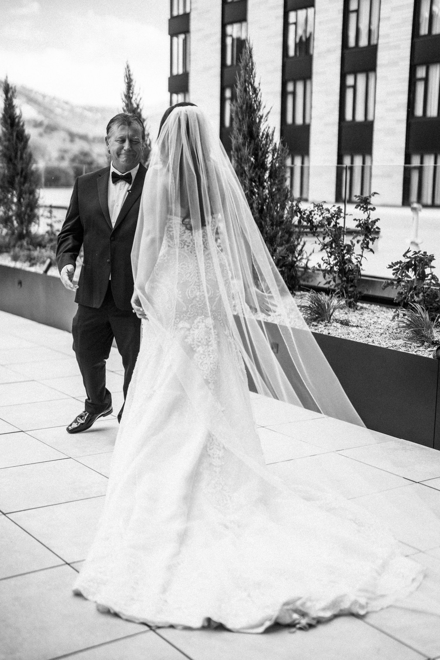 Black and white photo of father walking toward bride in lace gown and cathedral veil on rooftop.
