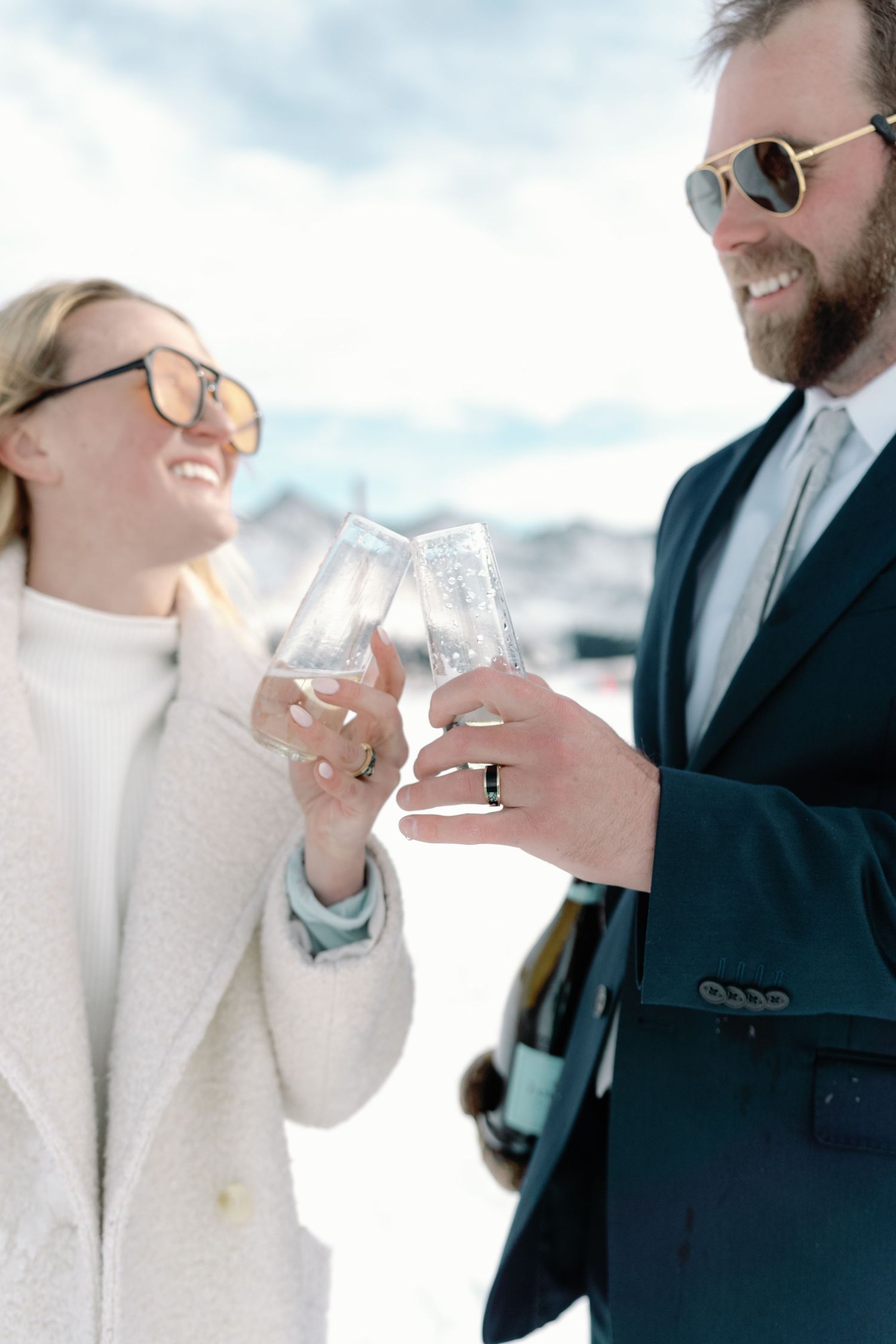 Couple in formal wear and sunglasses toasting champagne glasses together on a snowy mountain.