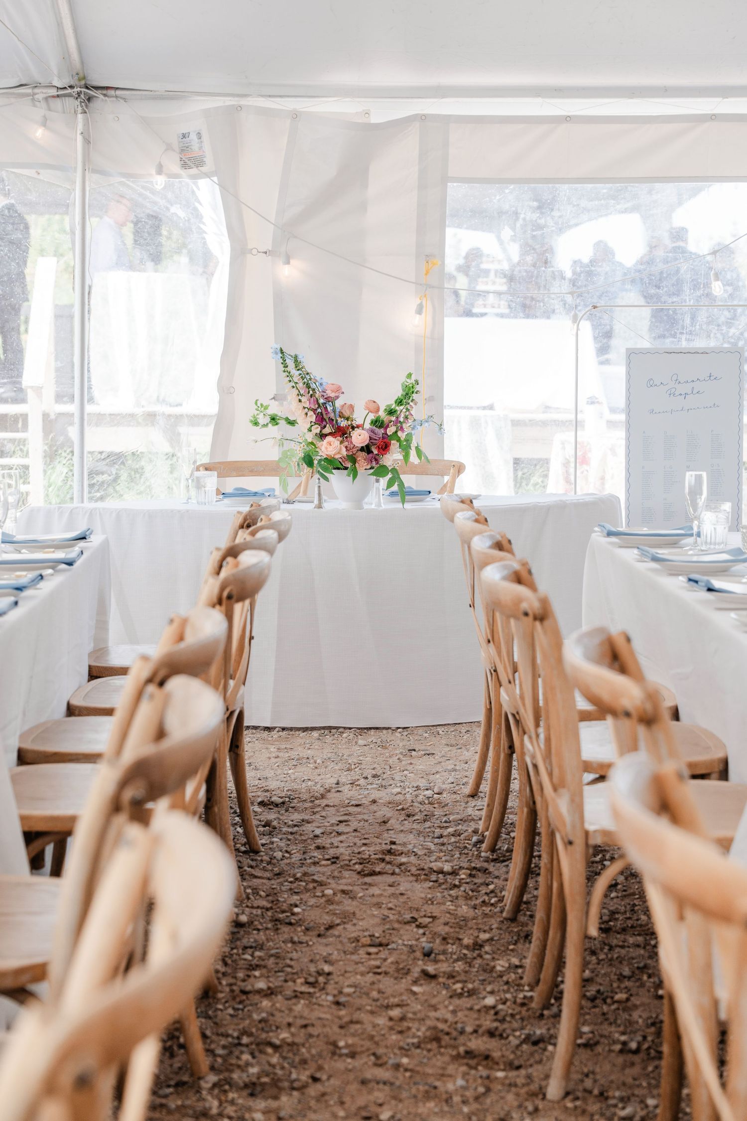 Elegant wedding reception tent with long white-draped tables, wooden crossback chairs, and a floral centerpiece.
