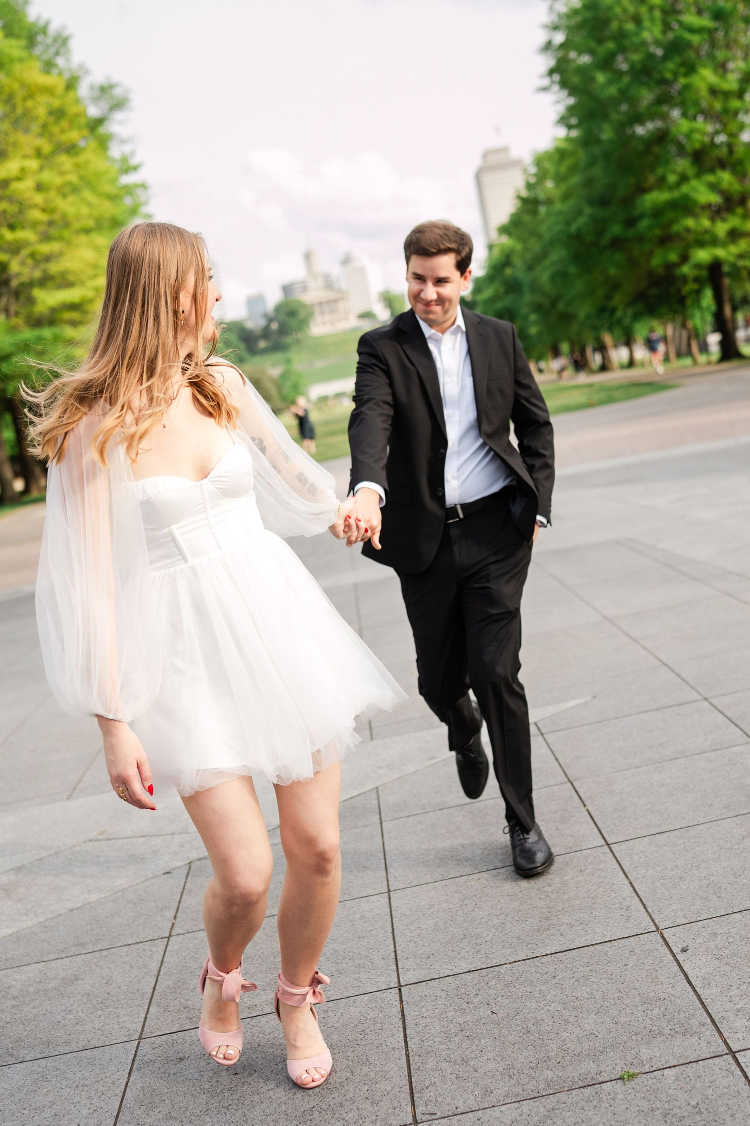 Bride in white mini dress and pink heels holding grooms hand while walking in an urban park.