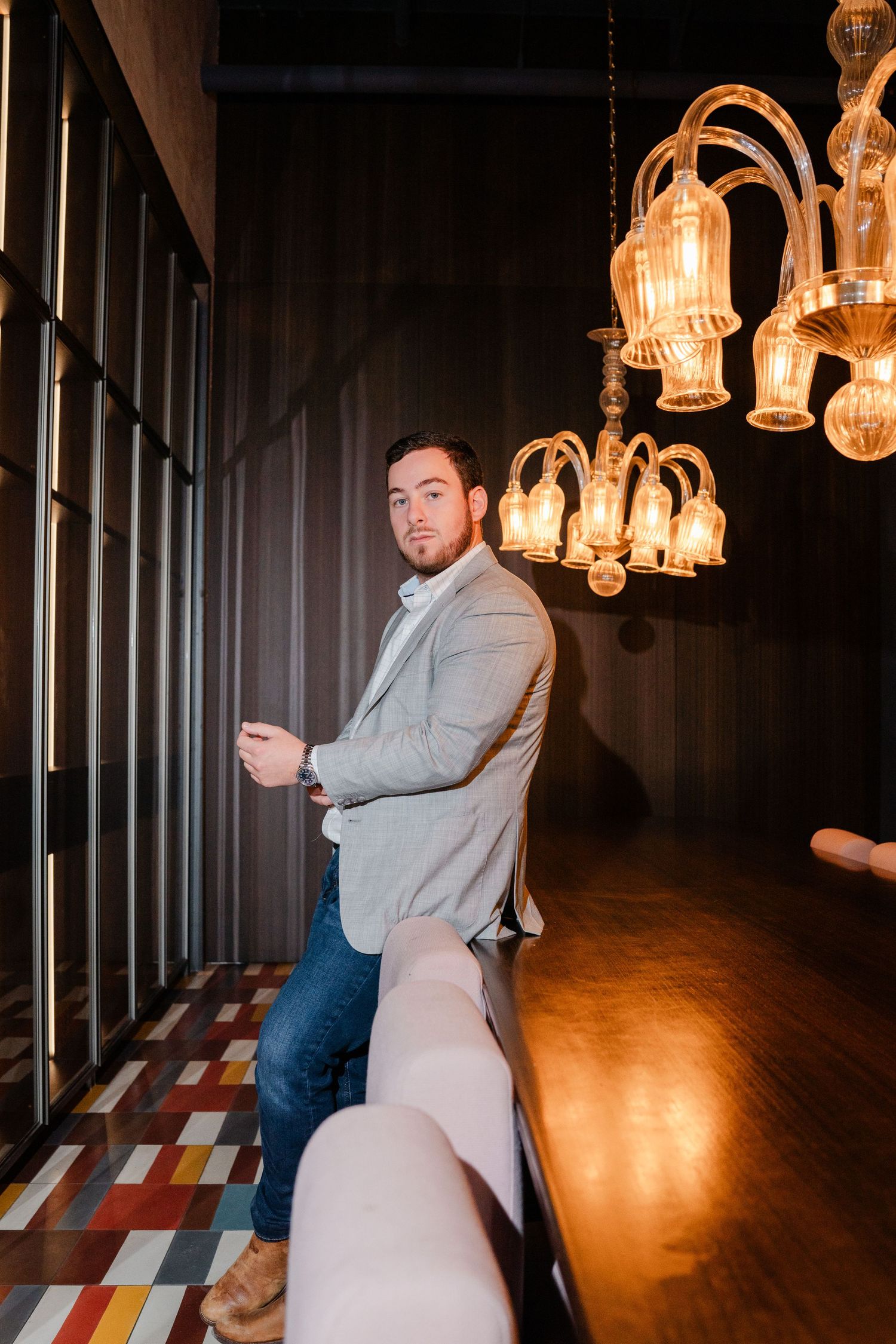 Handsome man in gray blazer leaning against bar counter under warm vintage chandelier lighting indoors.
