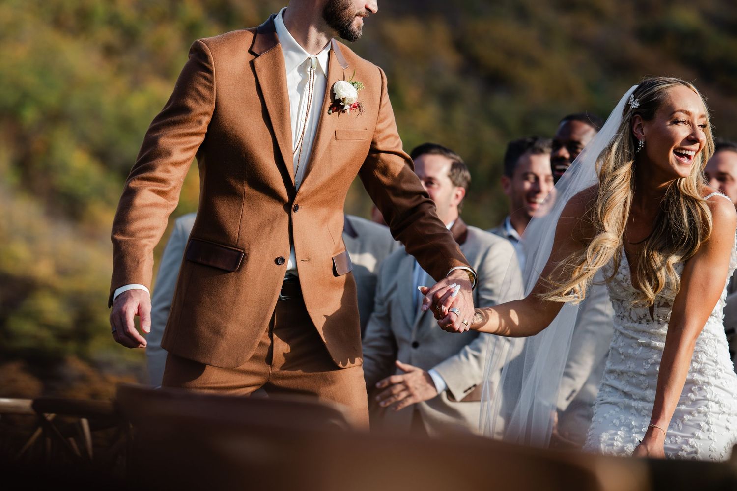 Joyful bride in white gown holds hands with groom in brown suit at outdoor wedding.