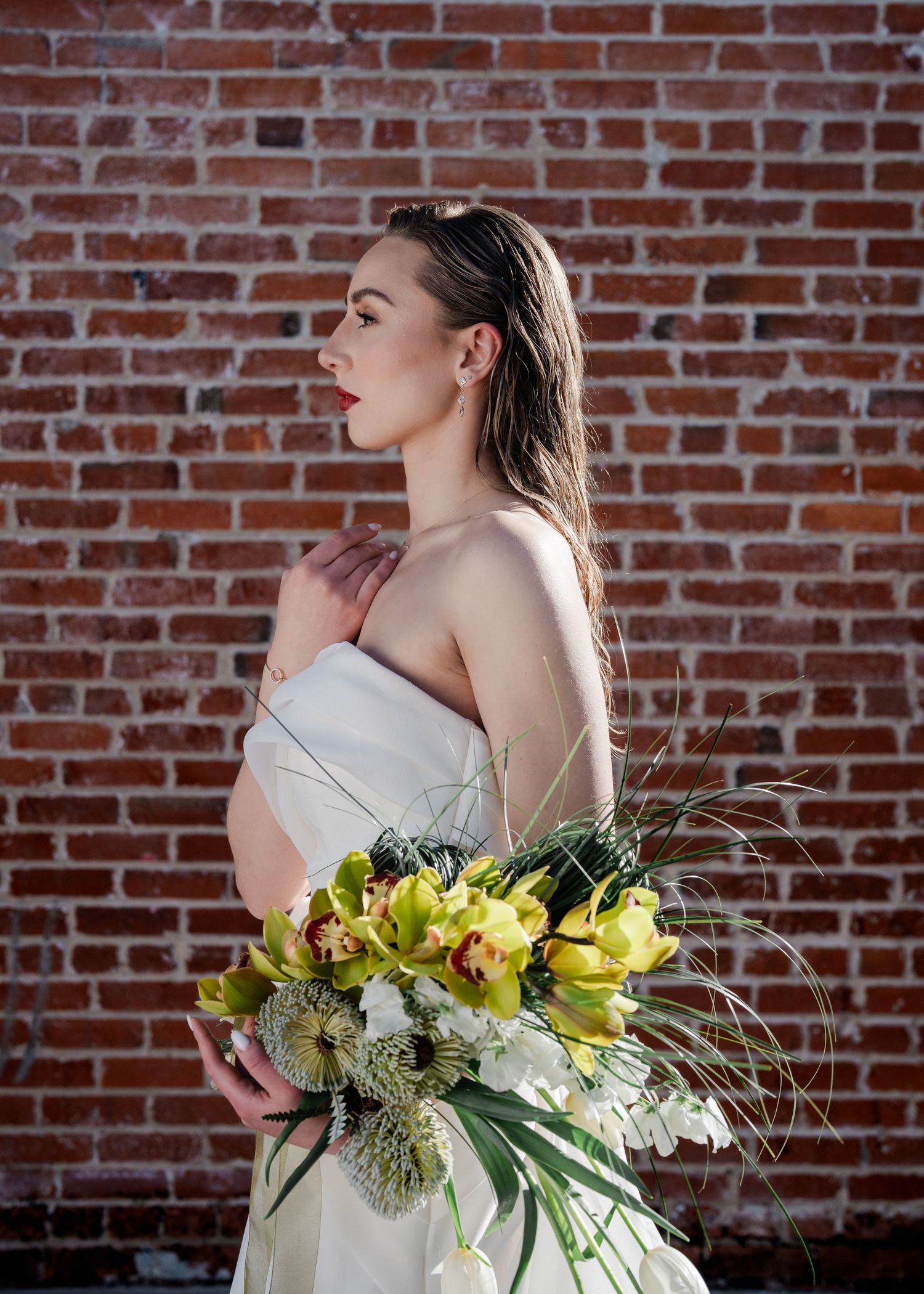 Elegant bride in a white strapless gown holds a tropical green orchid bouquet against a red brick wall.