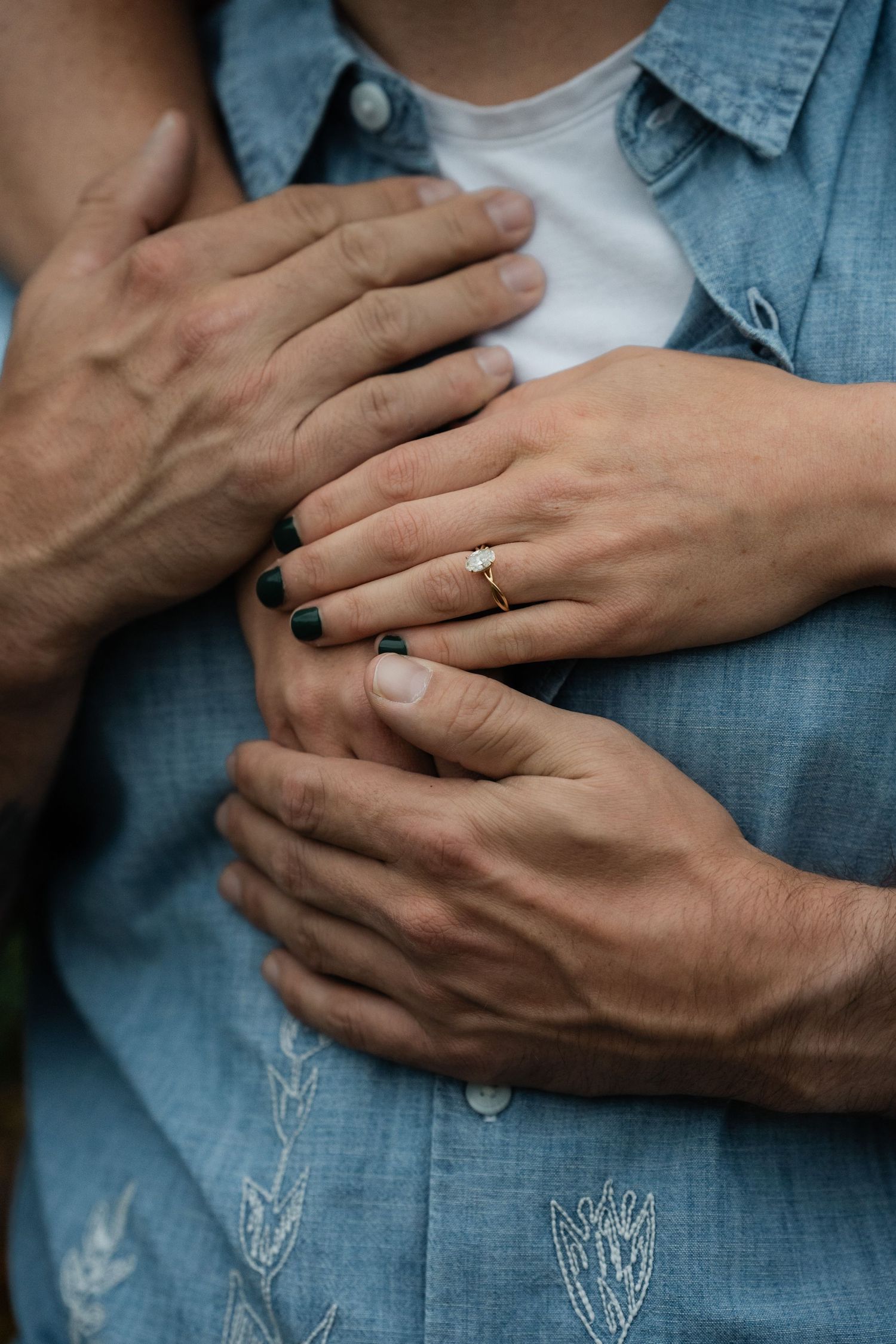 Close-up of couples hands showing diamond engagement ring, woman embracing man in blue chambray shirt.