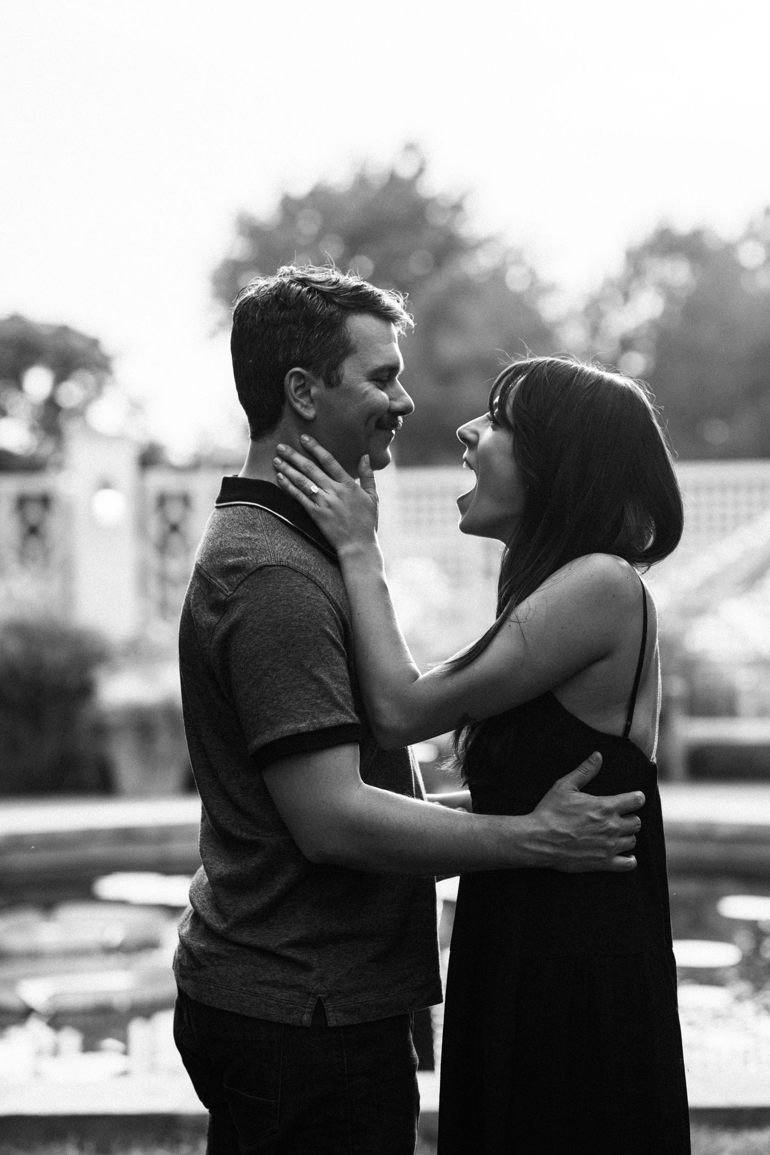 Black and white photo of couple embracing and laughing near garden fountain.
