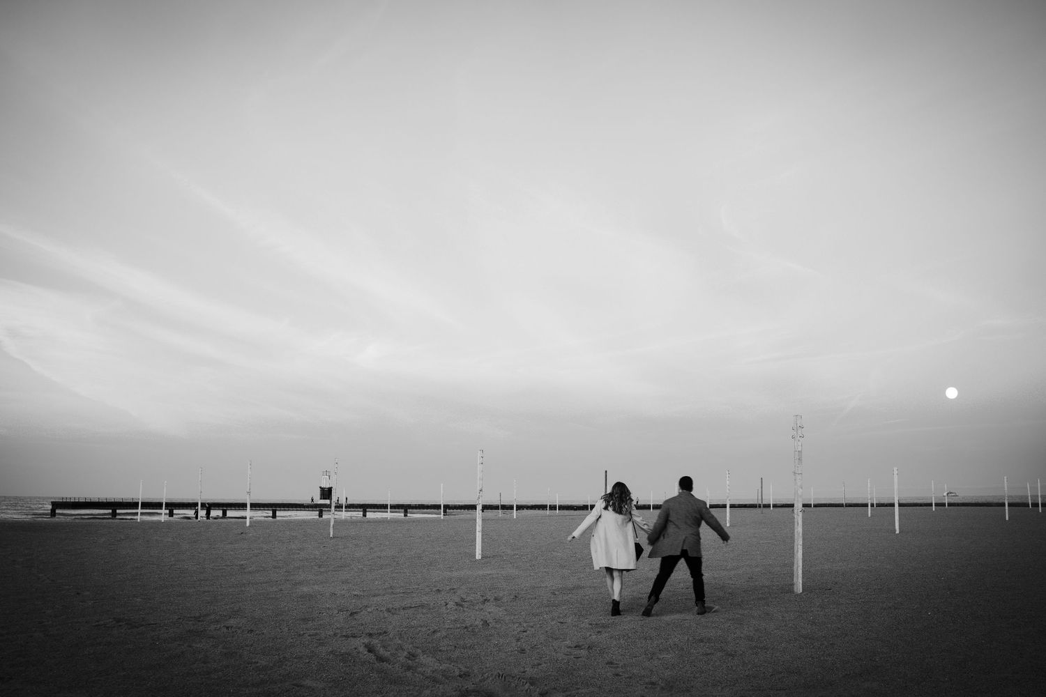 Black and white silhouette of couple holding hands walking on sandy beach at dusk with pier in background.