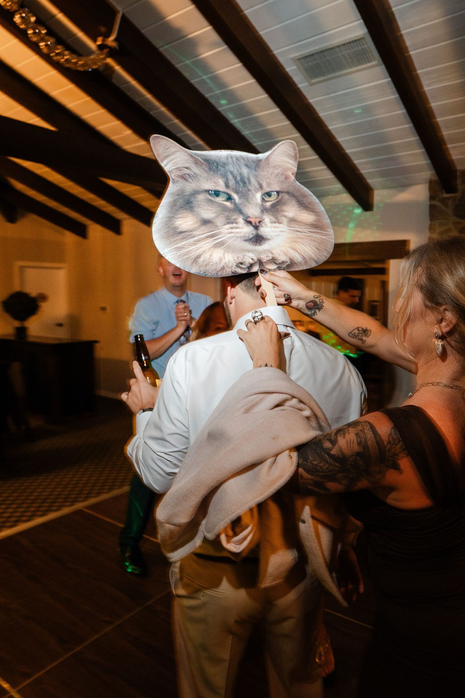 Wedding guests holding up a large decorative cat face prop at an indoor wedding reception celebration.