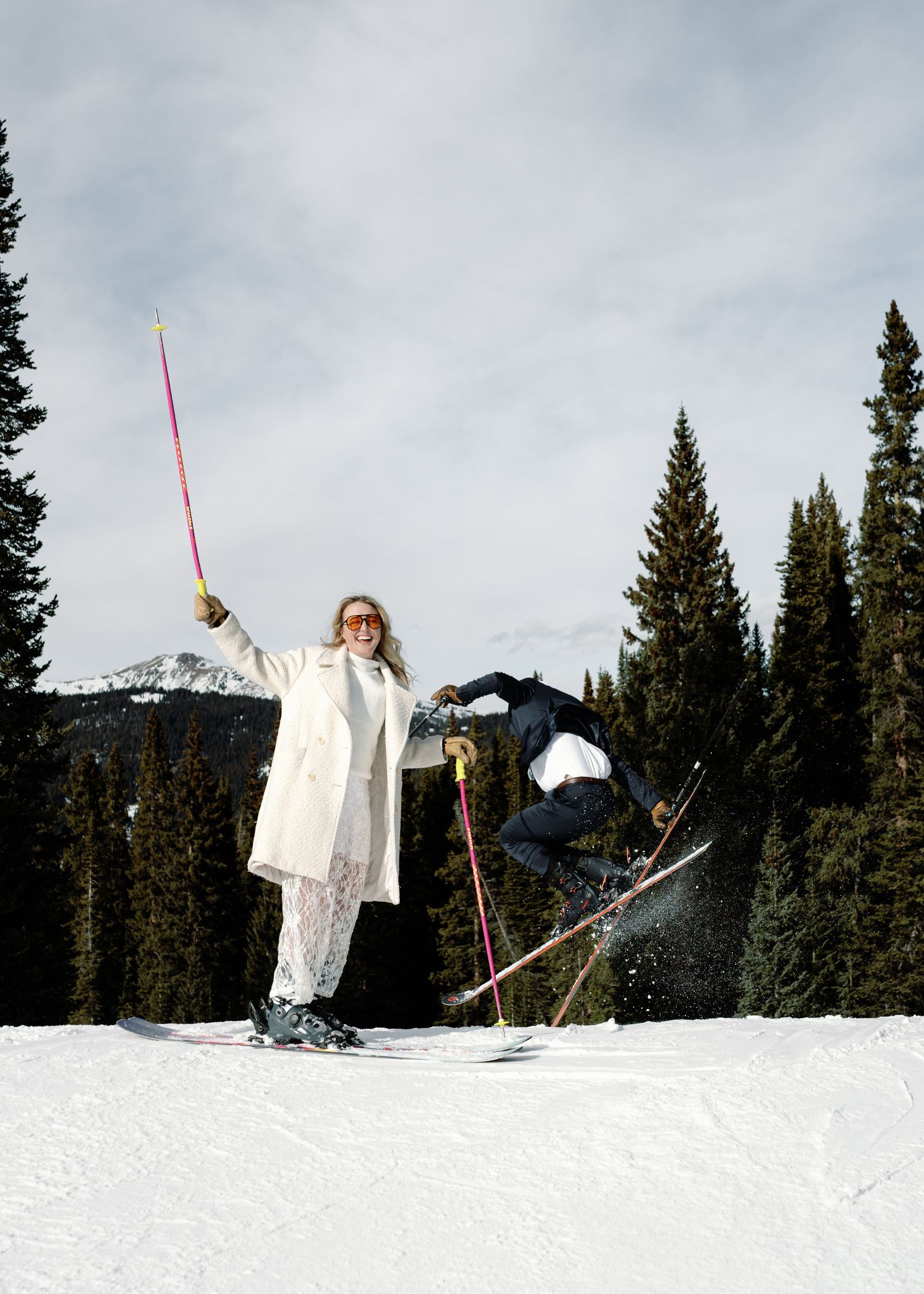 Woman in white coat raises pink ski pole as skier performs jump trick behind her on snowy slope.