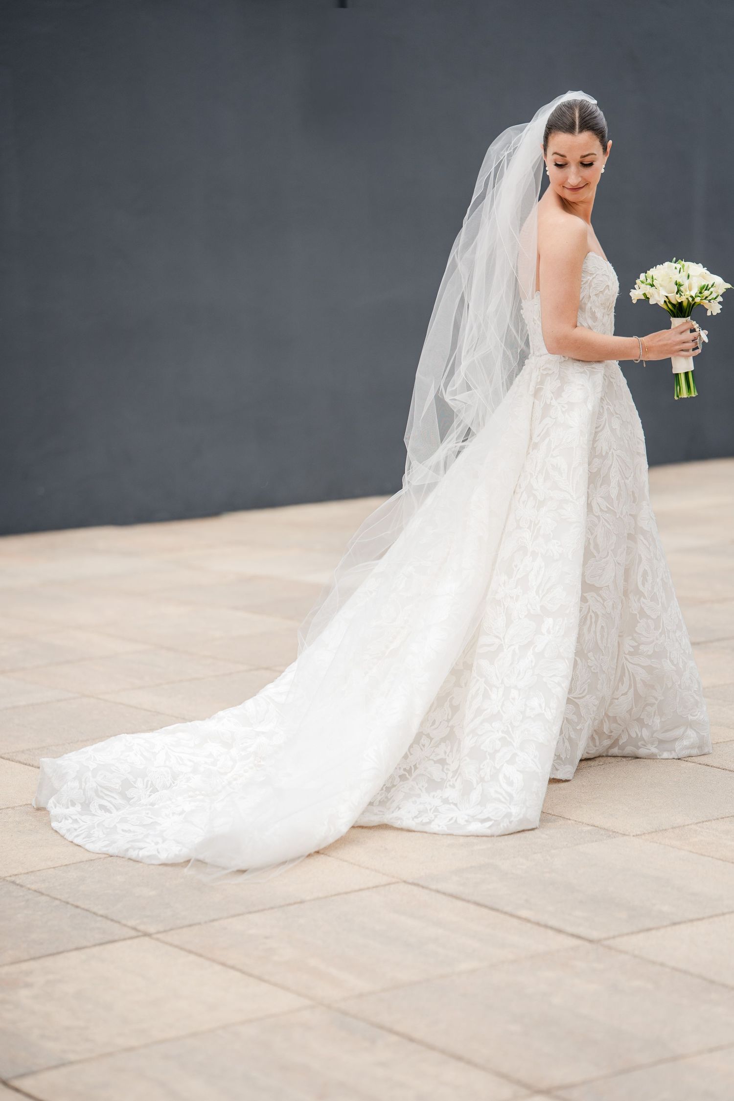 Elegant bride in lace ball gown with long veil holds white bouquet against a gray wall.