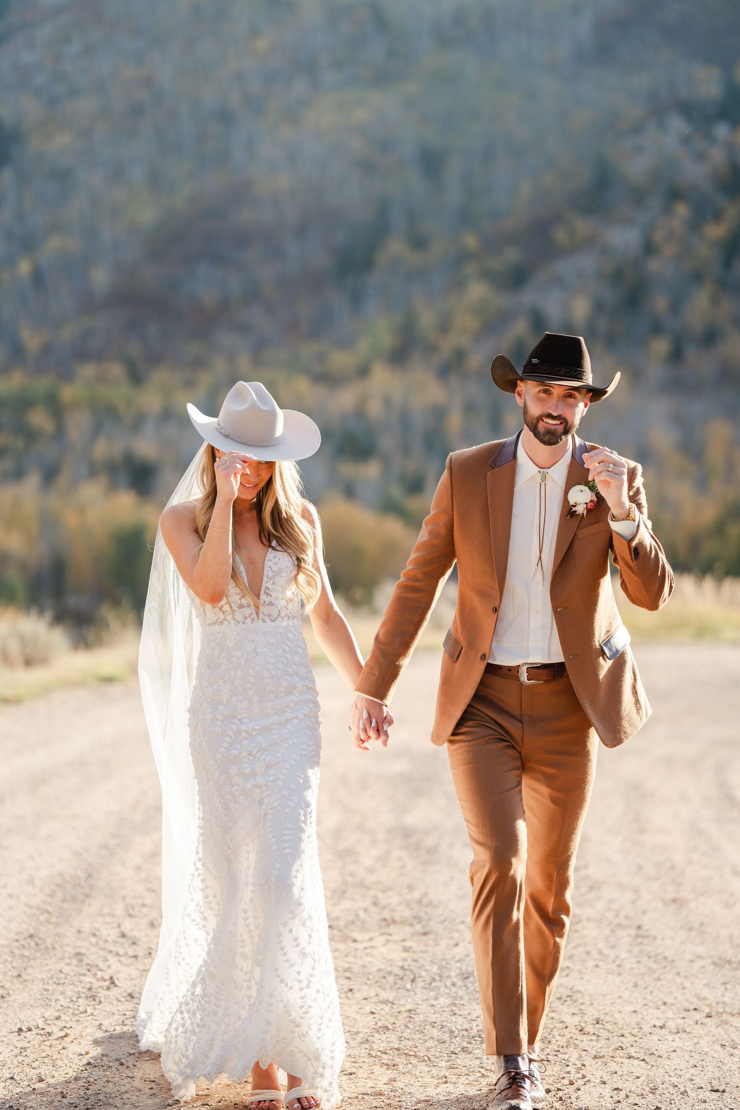 Bride and groom in cowboy hats holding hands walking on a dirt road with mountain backdrop.