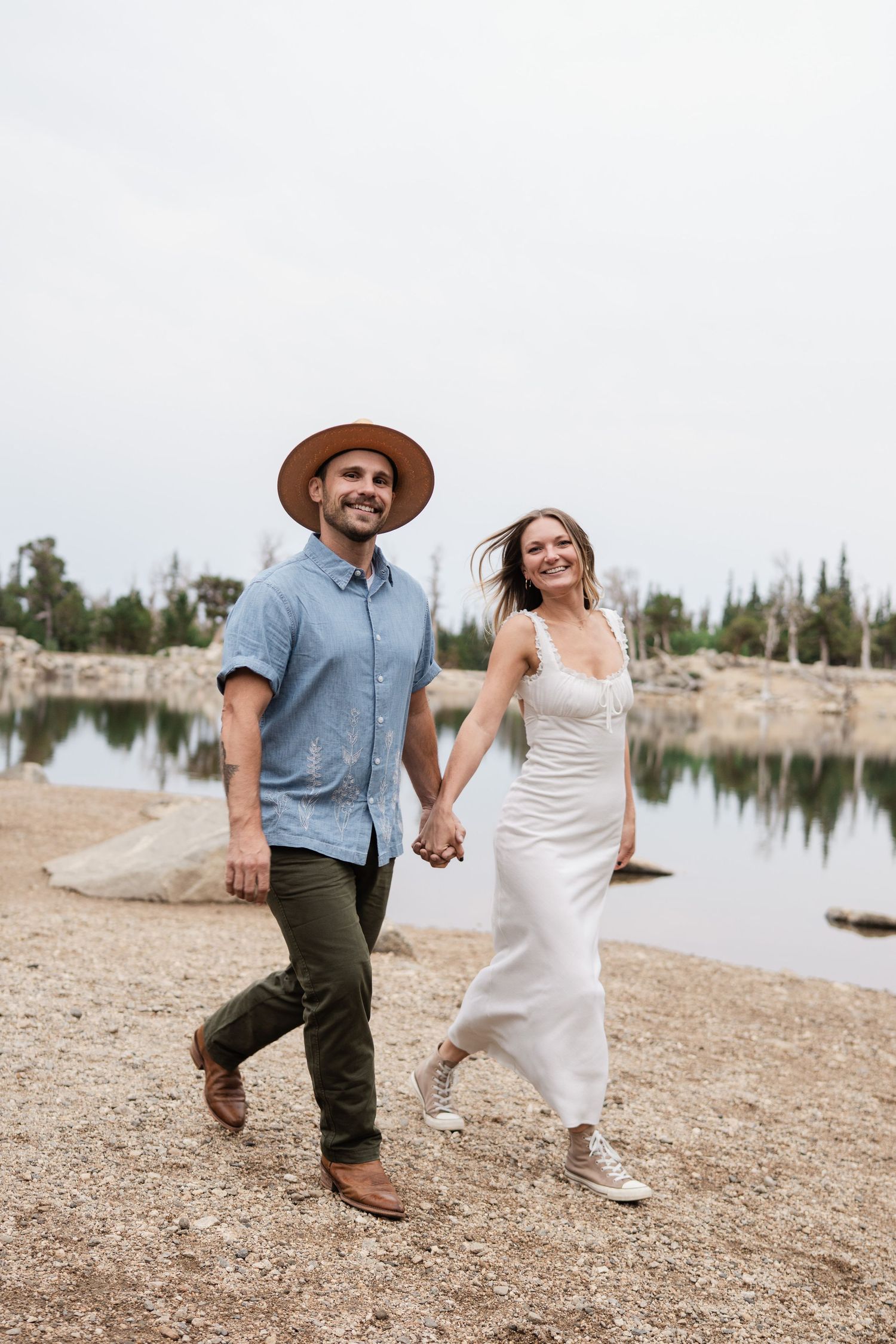 Happy couple holding hands walking near reflective alpine lake, man in hat and woman in white dress.
