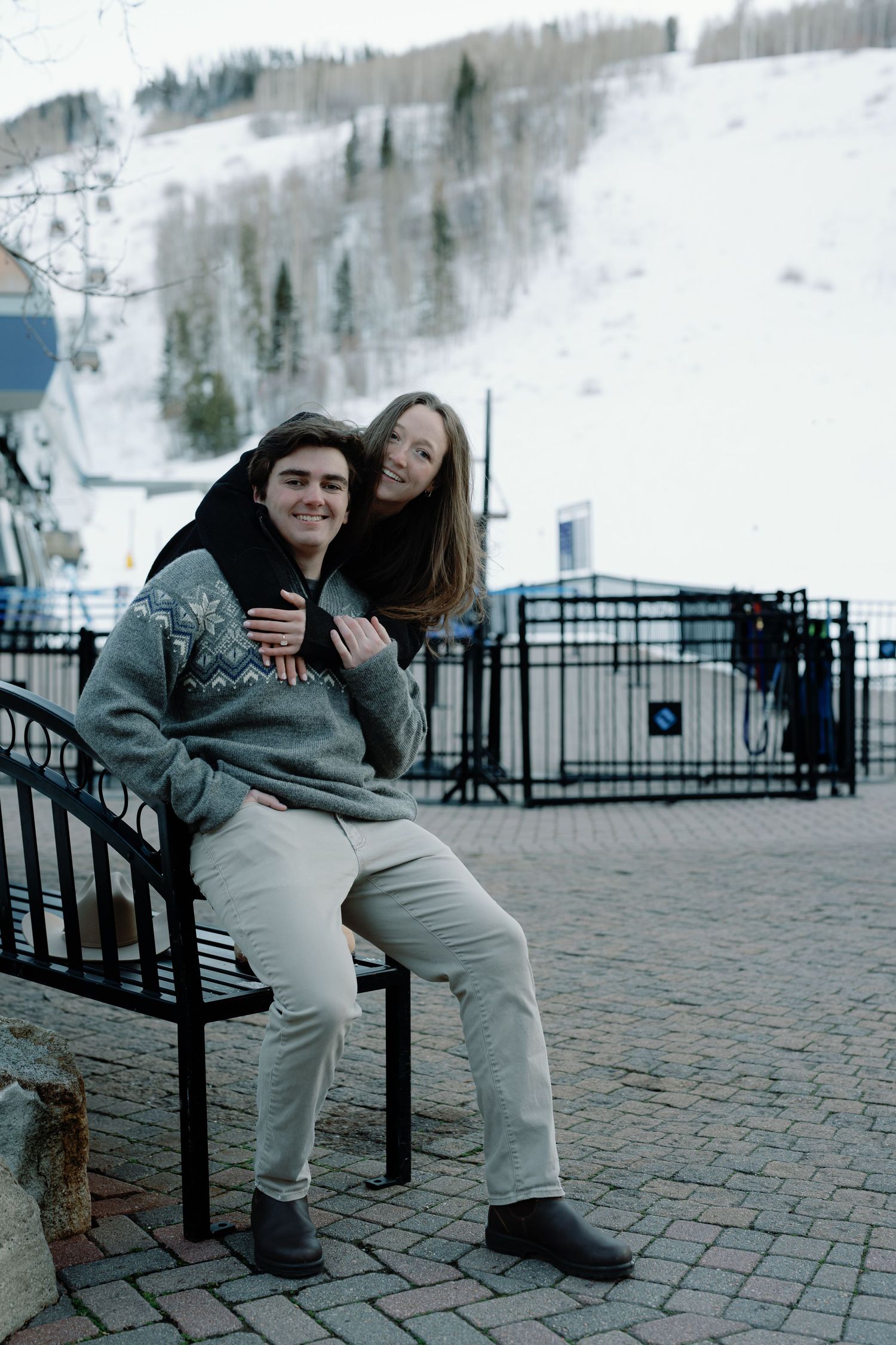 Smiling couple posing on a bench with a snowy ski mountain backdrop in a winter resort town.