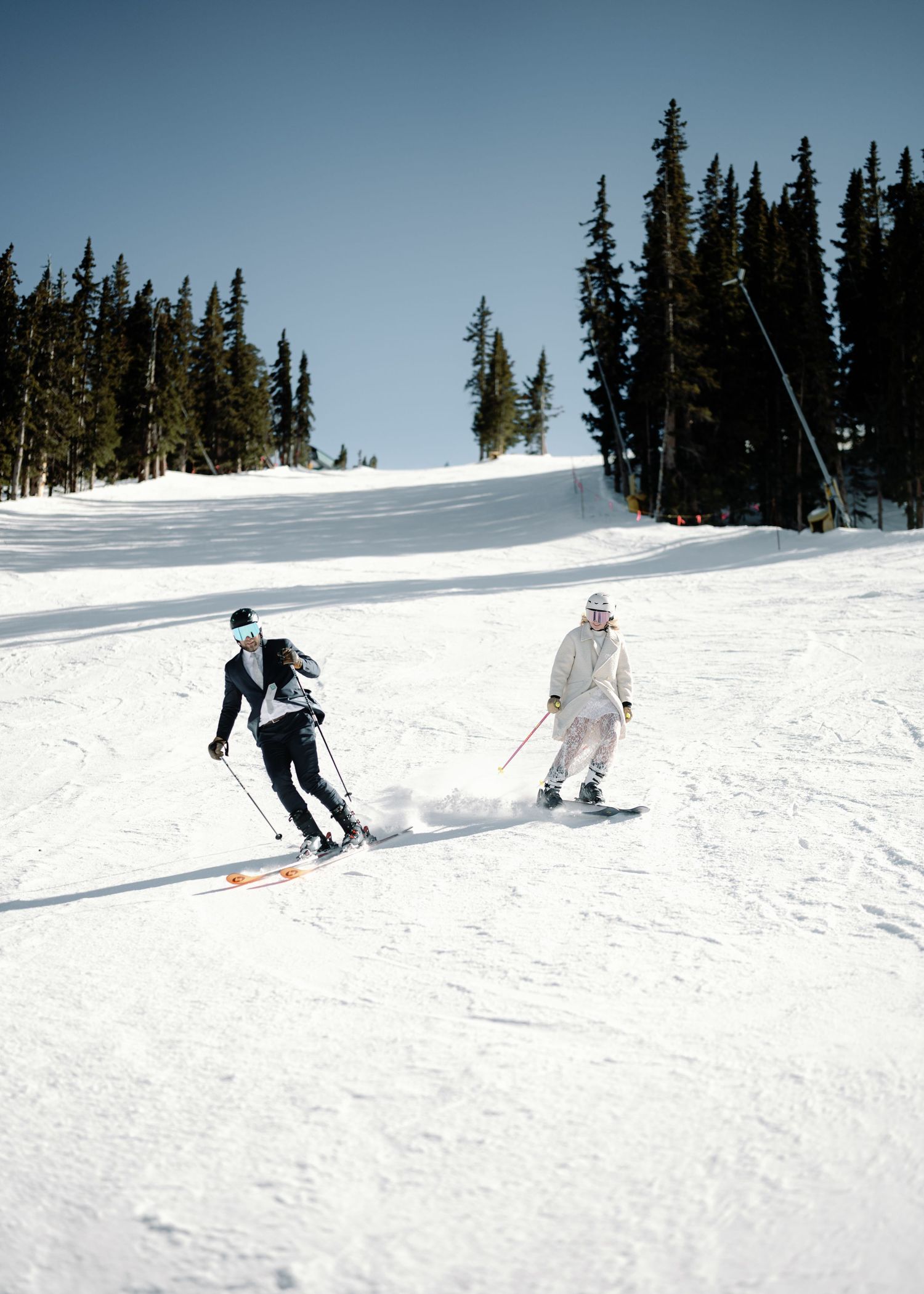 Bride and groom skiing down a snowy slope in wedding attire at a mountain resort.