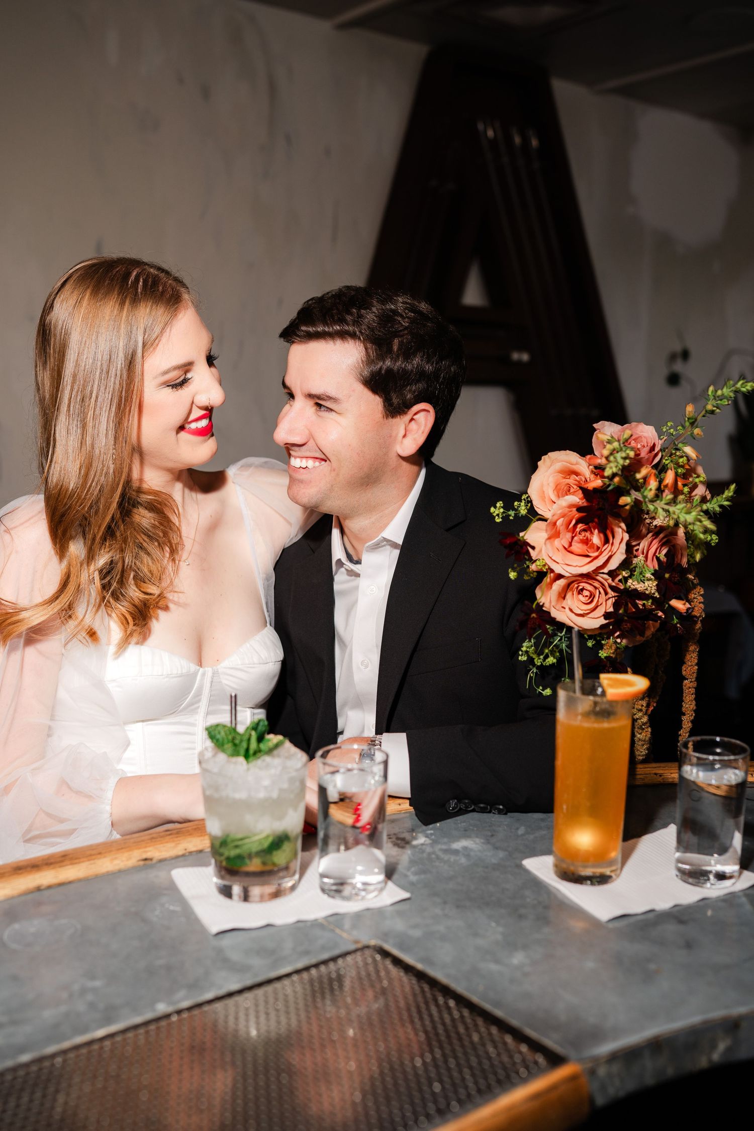 Smiling couple seated at a bar table with cocktails and a coral rose floral arrangement nearby.