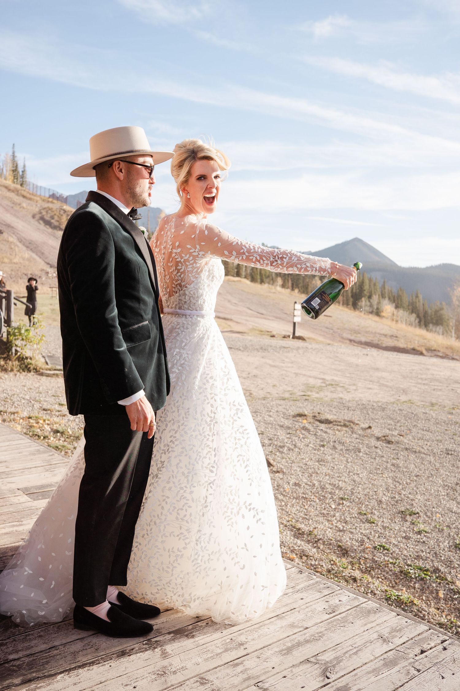 Laughing bride in a sparkling gown popping champagne with groom wearing a hat on a mountain boardwalk.