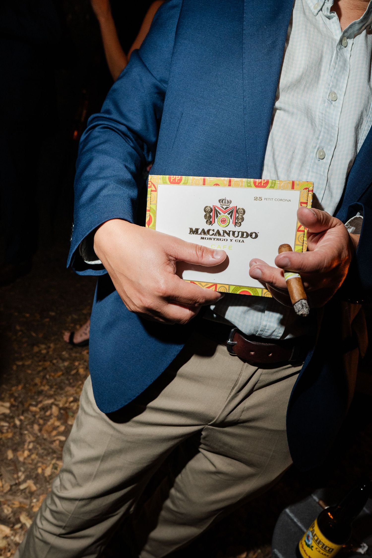 Wedding guest holds Macanudo cigar box and lit cigar at outdoor evening reception.