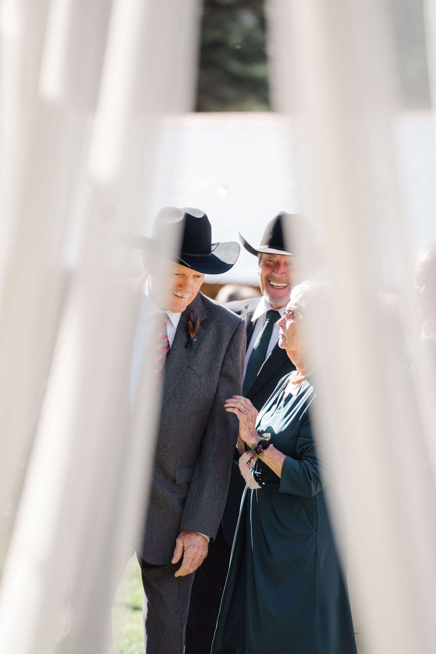 Two men in suits and black cowboy hats laughing together at an outdoor wedding event.