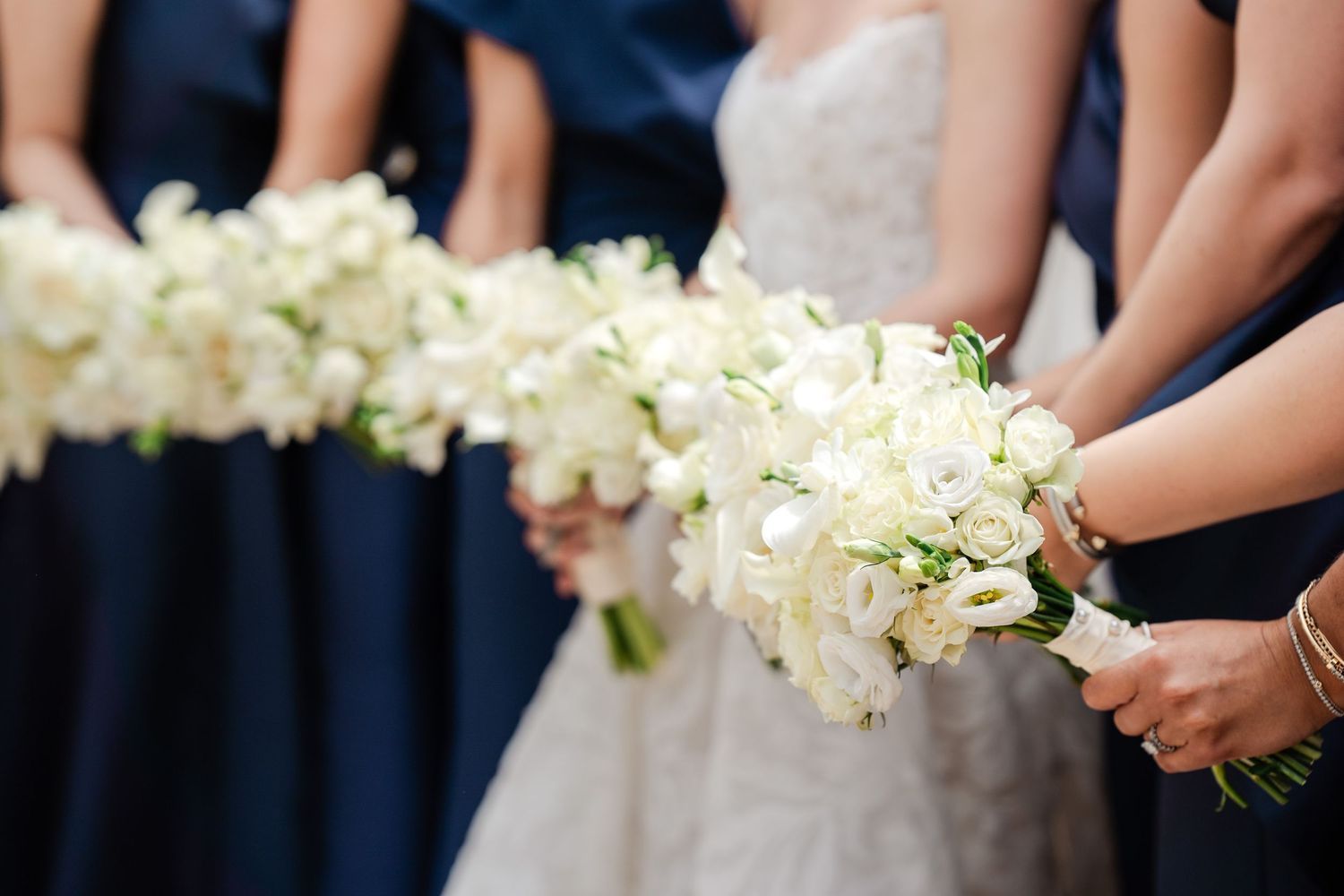 Bridesmaids in navy blue dresses holding white rose bouquets alongside bride in lace wedding gown.