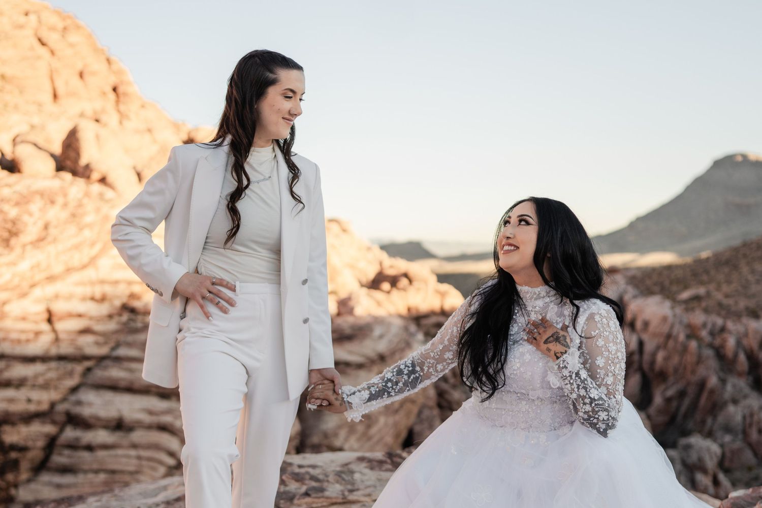 Two brides in white attire hold hands and smile at each other against a stunning red rock canyon backdrop.