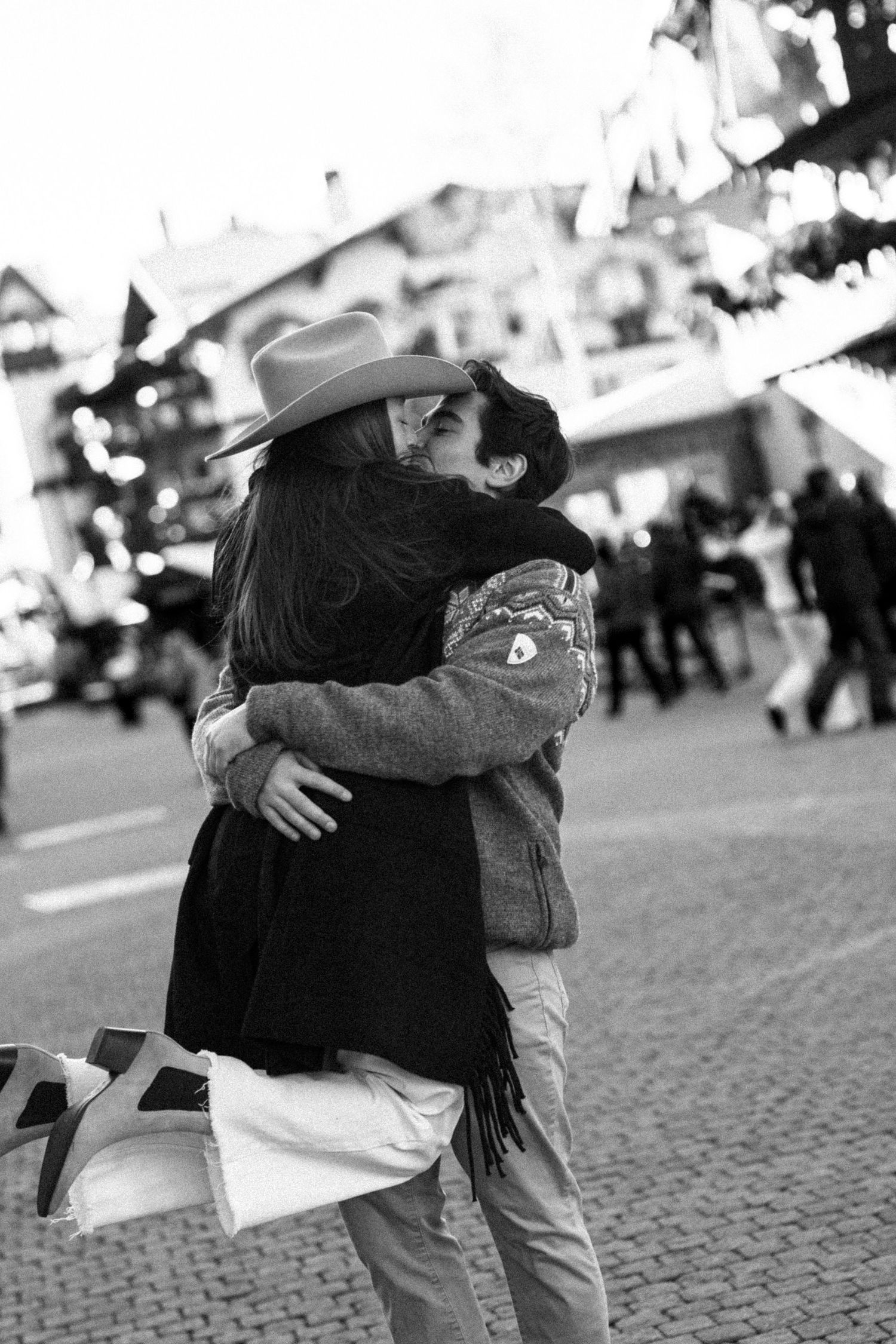 Black and white photo of couple embracing and kissing in a lively winter town square.