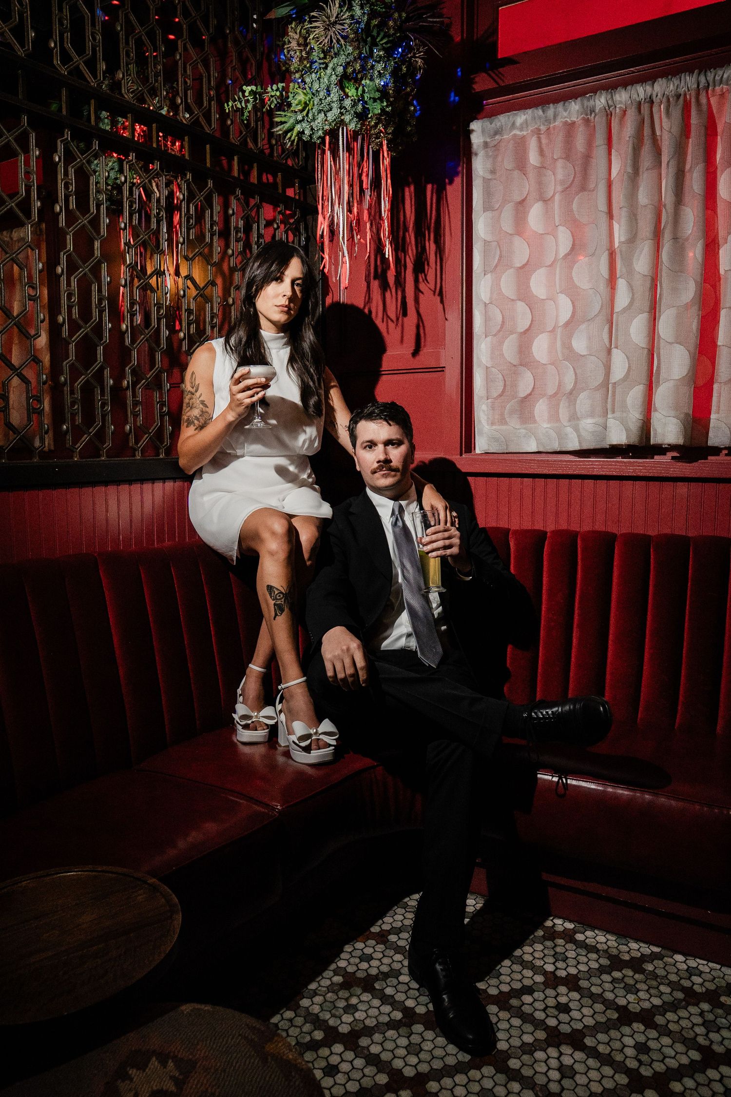 Couple in wedding attire relaxing in a moody red velvet booth at an upscale vintage bar.