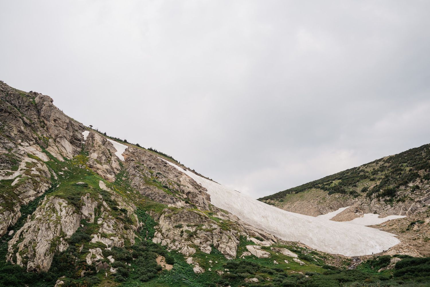 Snow patch on rocky mountain slope with green alpine vegetation under overcast sky.