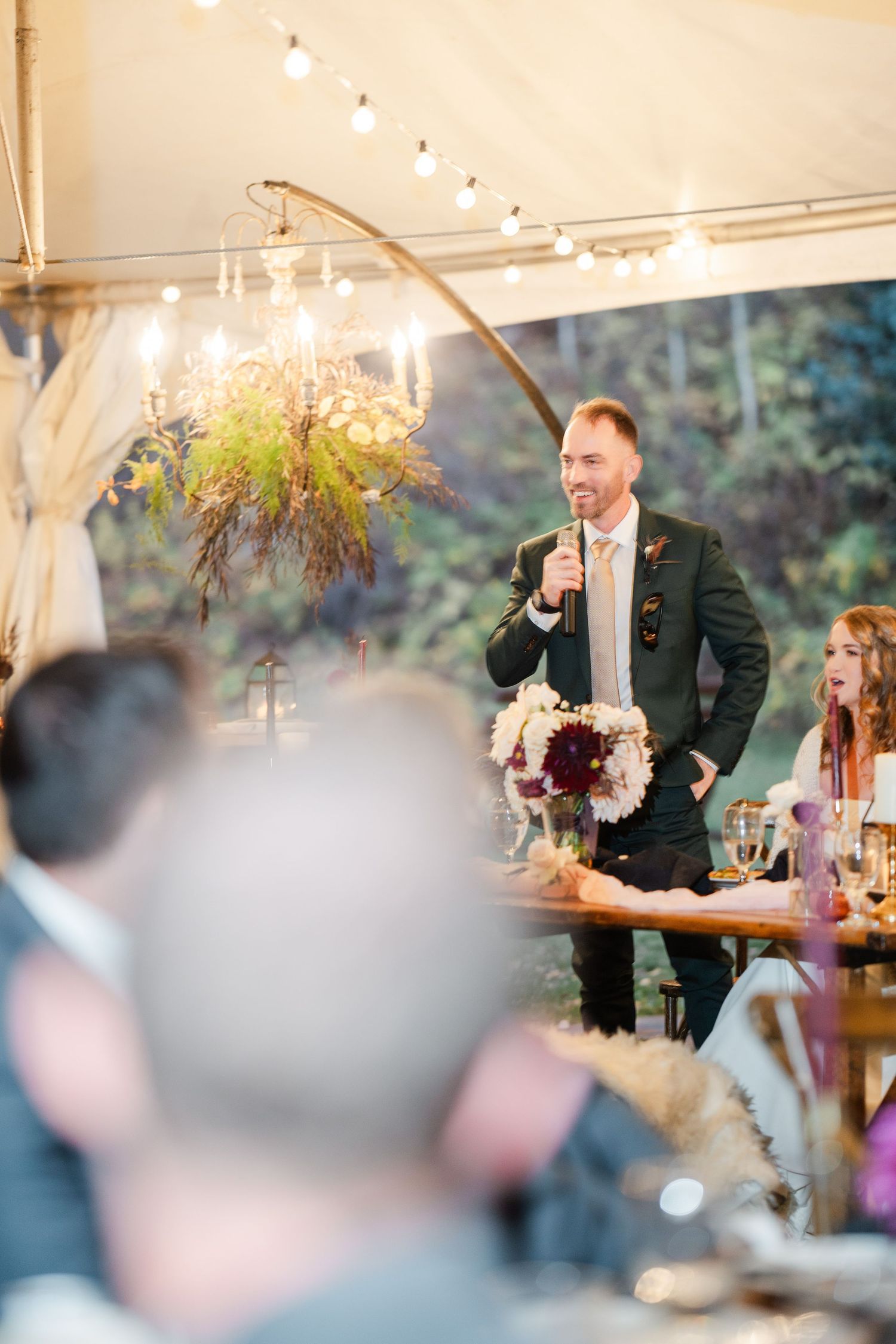 Smiling best man giving a toast with microphone at an elegant outdoor tented wedding reception.