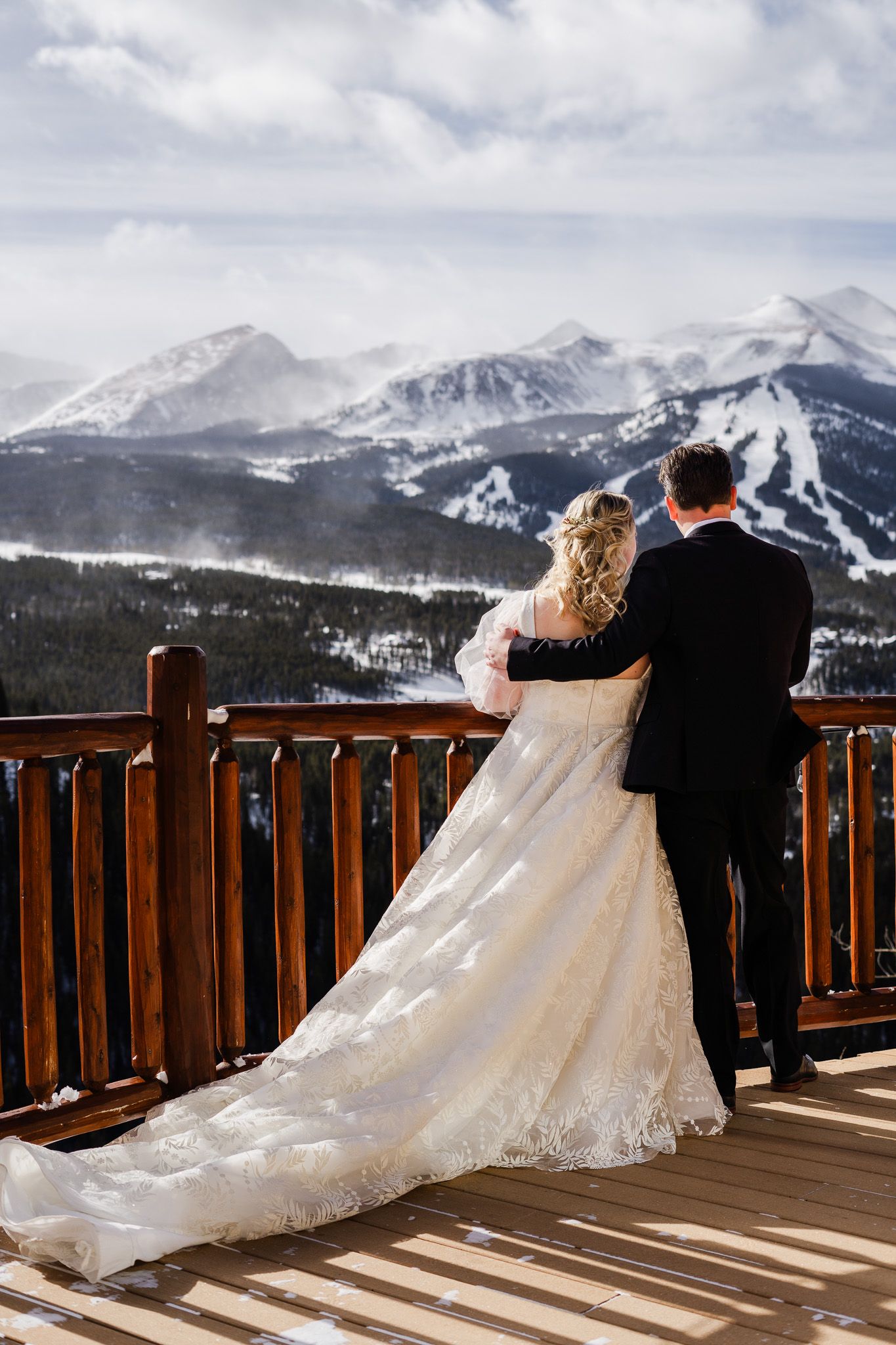 Bride and groom embrace on a wooden deck overlooking snow-covered ski resort mountains on their wedding day.