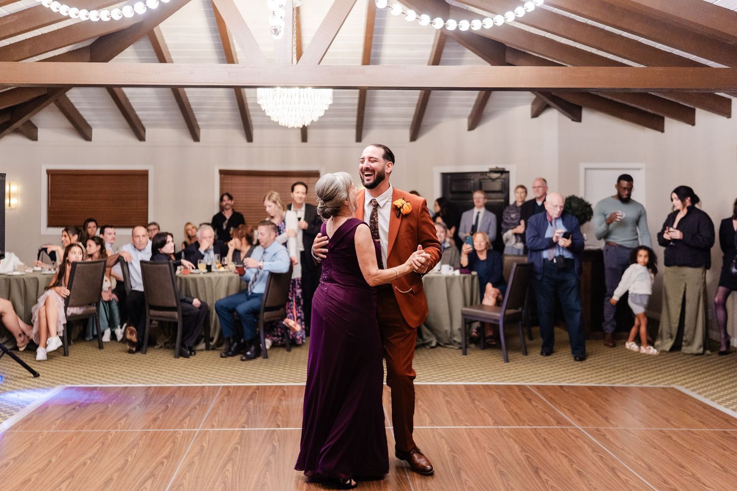 Groom in rust suit shares mother-son dance at elegant wedding reception venue.