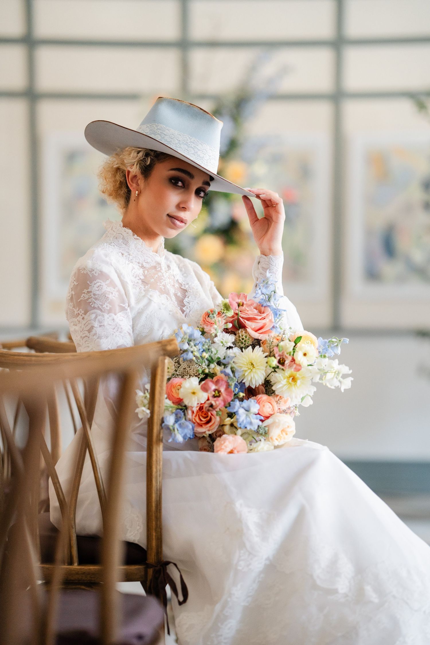 Stylish bride in lace gown and silver wide-brim hat holds a pastel floral bouquet while seated.