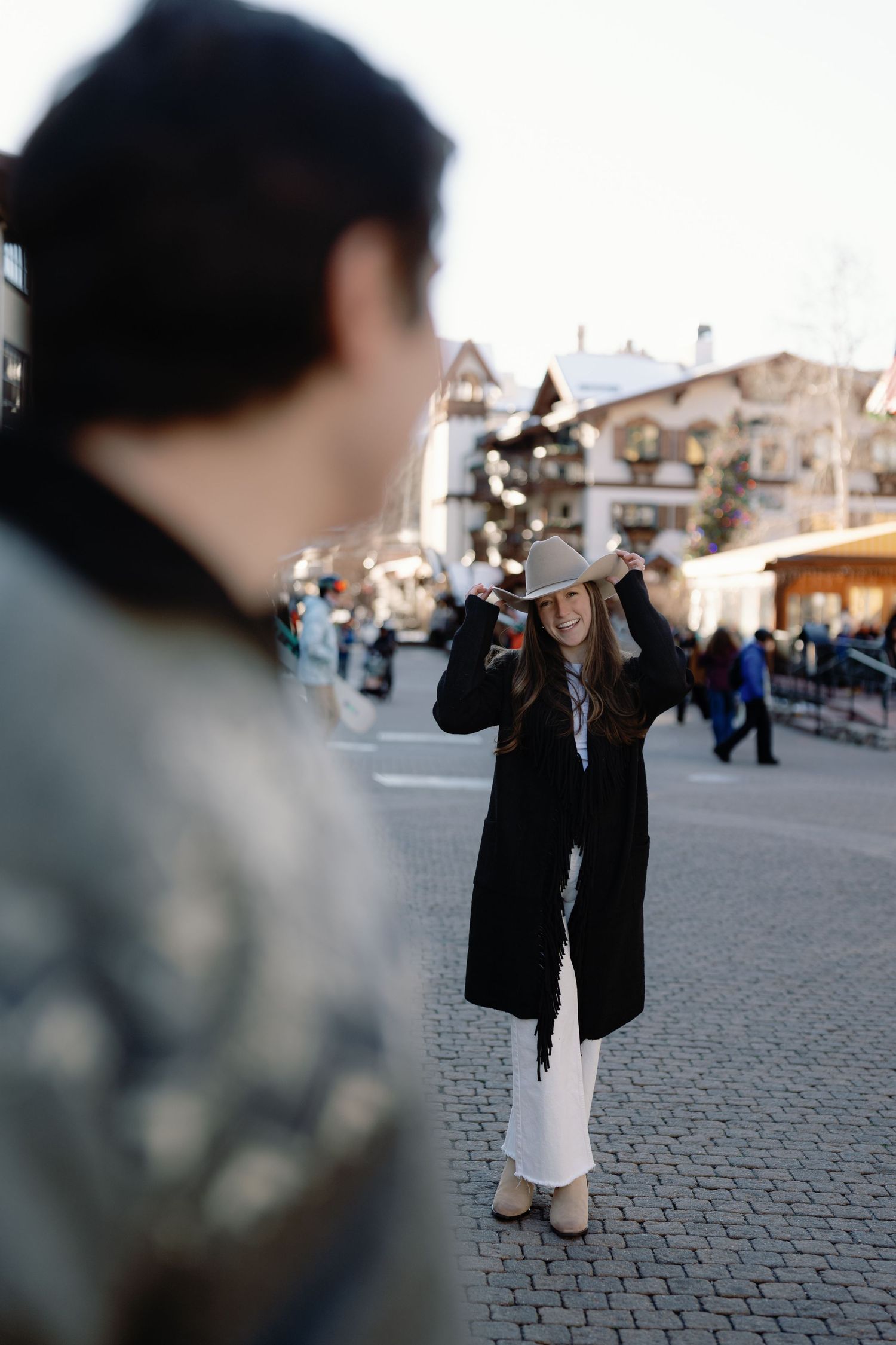 Smiling woman in cowboy hat and black coat posing in a festive snowy mountain village.