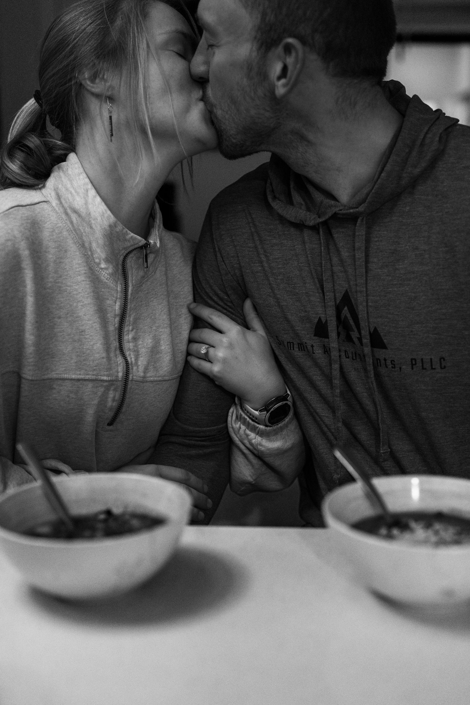 Black and white photo of couple sharing a kiss over bowls of soup at a kitchen table.